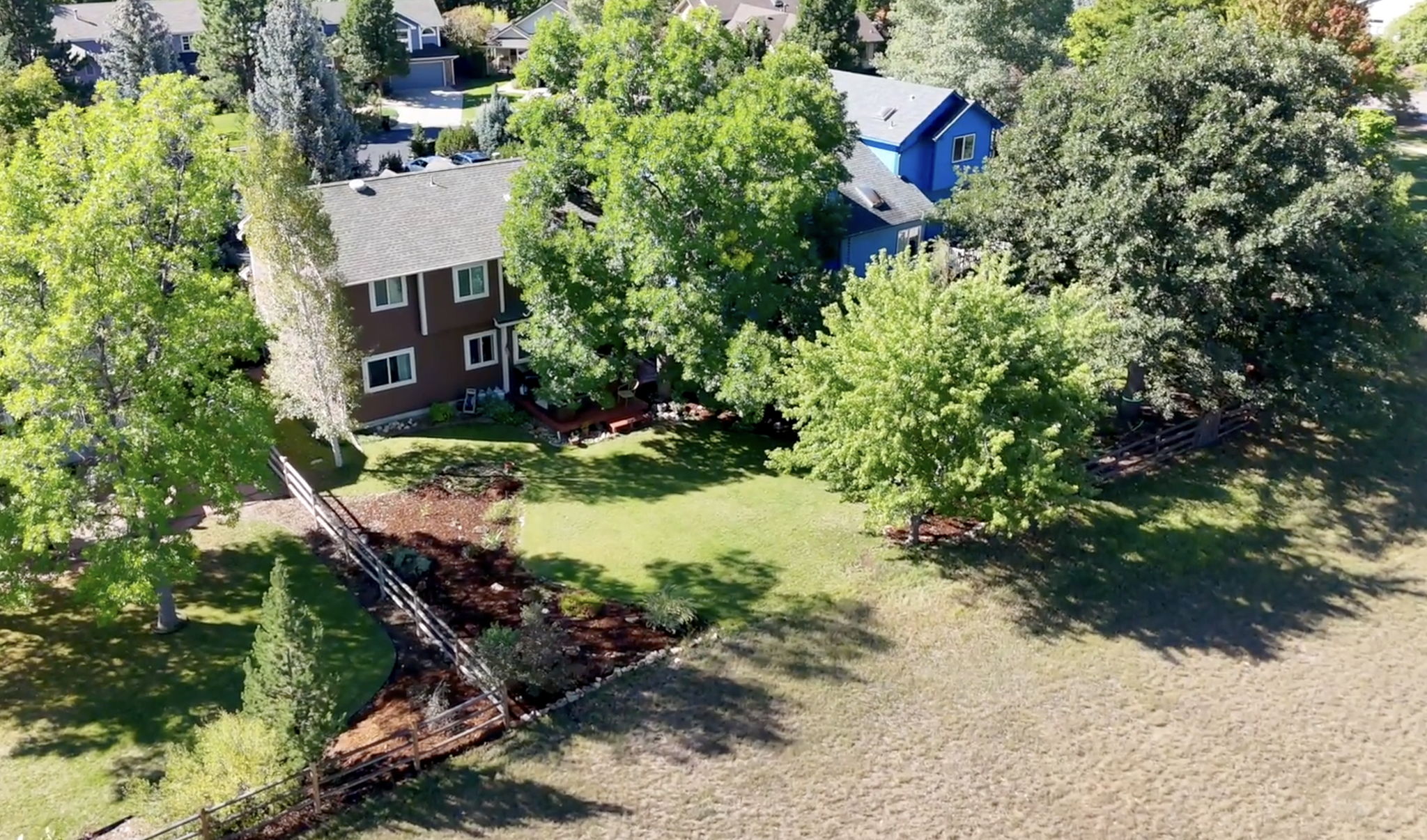 This aerial view showcases a well-maintained property with a brown two-story house, a blue house, mature trees, and a spacious green lawn. A wooden fence borders the yard, adding a rustic touch. The overall impression is one of a peaceful and private residential setting.