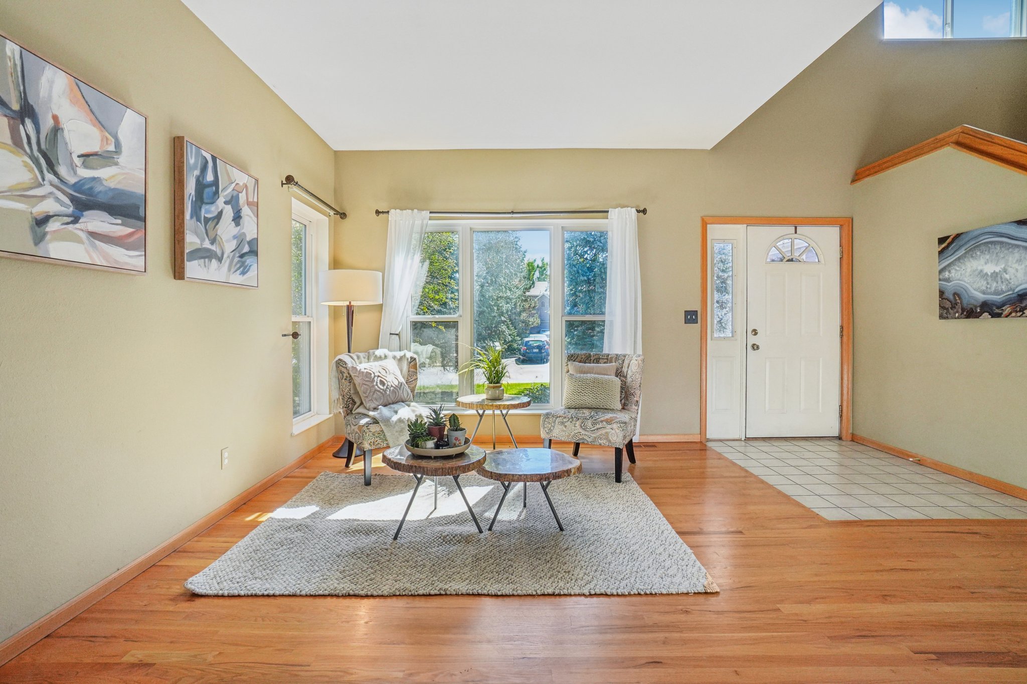 This inviting living room features hardwood floors, a neutral color palette, and ample natural light streaming through a large window. Two patterned armchairs flank a set of rustic coffee tables, creating a cozy conversation area. Artwork adorns the walls, adding a touch of personality to the space.