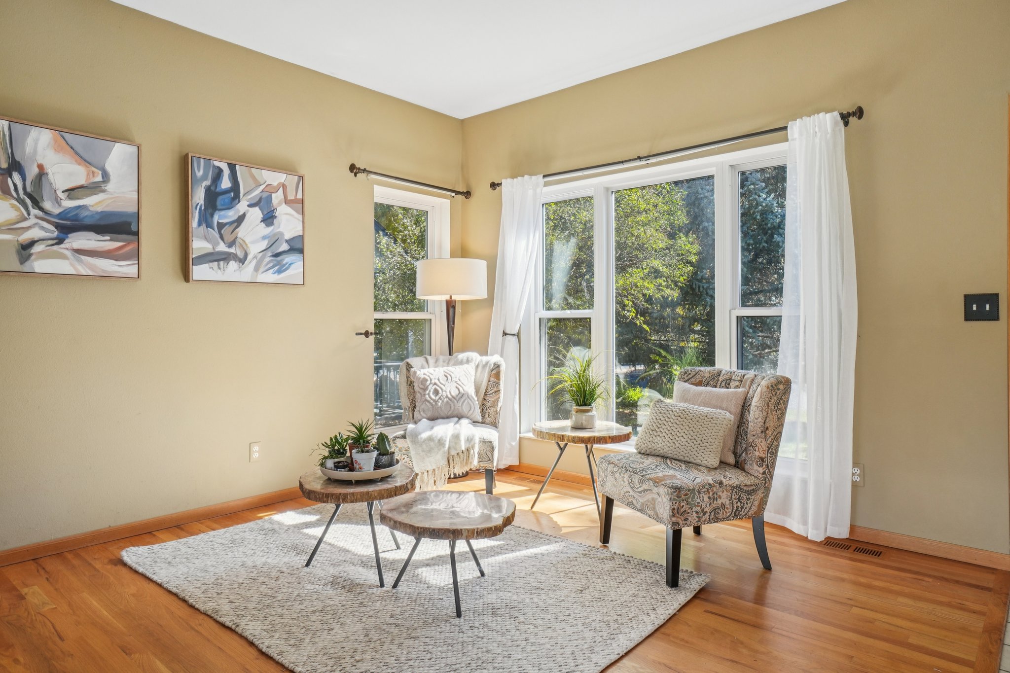 This inviting living room features two patterned armchairs flanking a large window with sheer white curtains, allowing natural light to fill the space. Two small, rustic-style side tables and a neutral-toned area rug add to the cozy atmosphere. Abstract artwork adorns the walls, complementing the warm, neutral color palette of the room.