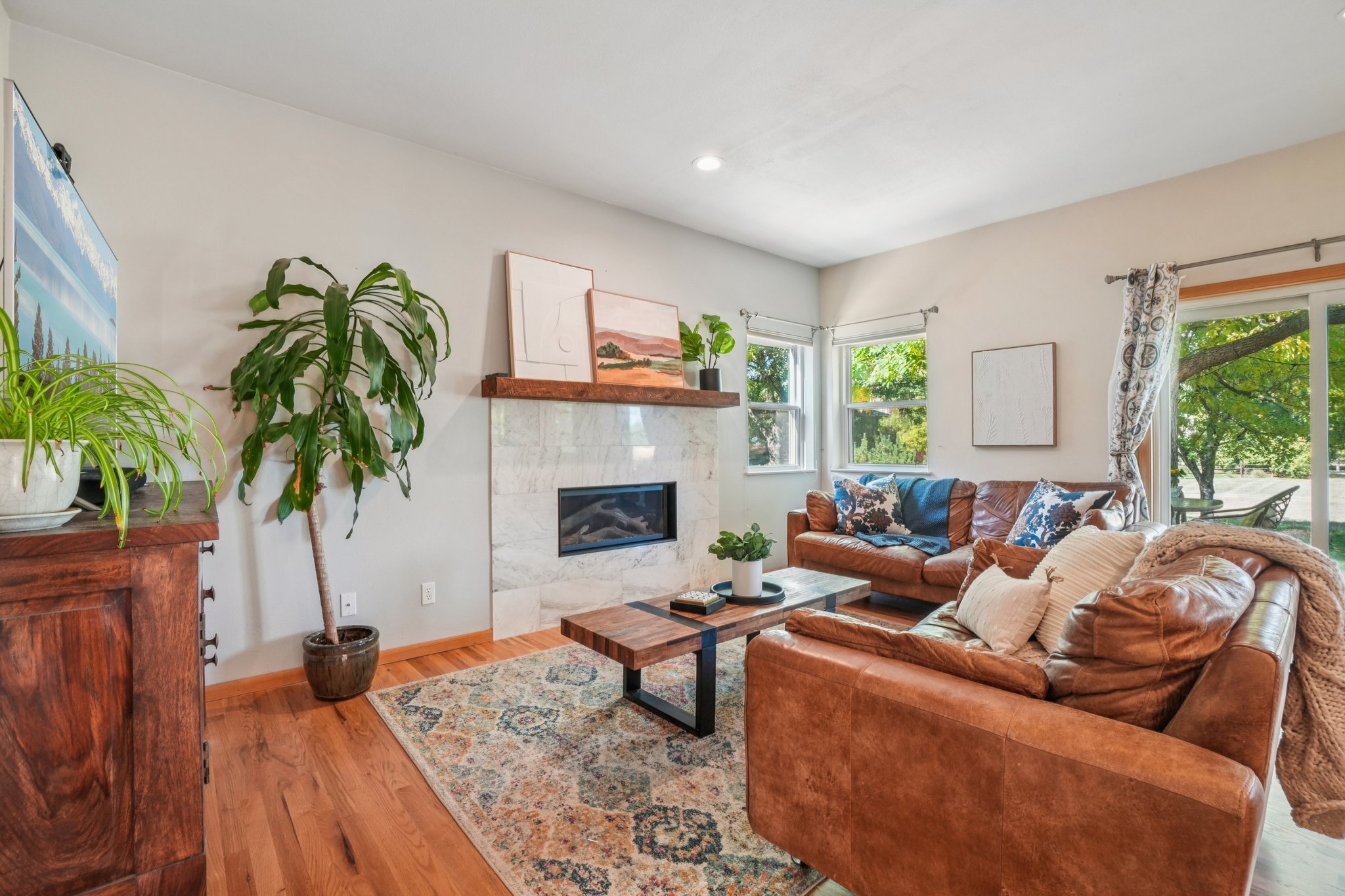 This is an interior shot of a living room featuring hardwood floors, a marble fireplace with a wooden mantel, and two brown leather sofas. A patterned area rug sits in the center of the room, and natural light streams in through the windows and sliding glass door. The room is decorated with plants and artwork, creating a warm and inviting atmosphere.