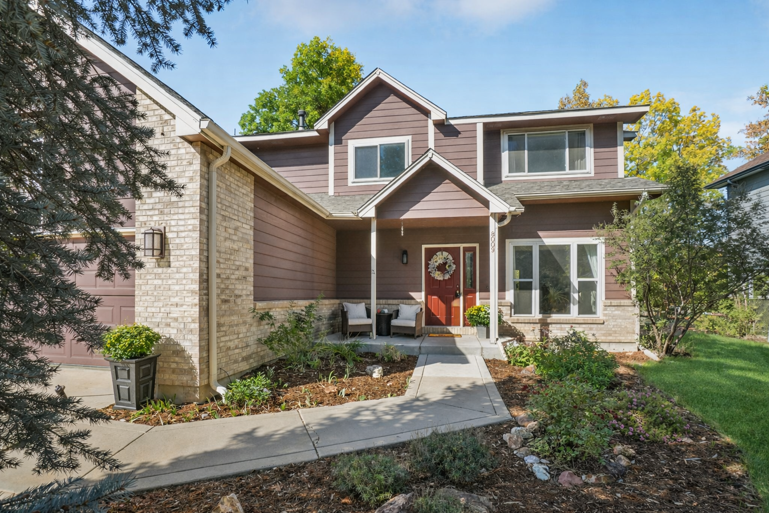 This is a front exterior view of a two-story home with a well-maintained yard. The house features a combination of brick and siding, with a covered entryway and a red front door adorned with a wreath. Landscaping includes mature trees, shrubs, and a concrete walkway leading to the entrance, creating a welcoming and established curb appeal.