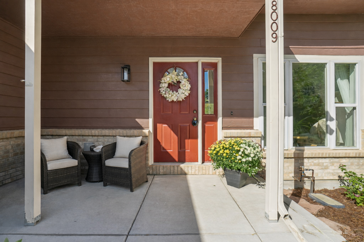 This is a welcoming entryway featuring a red front door adorned with a decorative wreath. Two wicker chairs with light cushions flank a small table, creating a cozy seating area. The exterior is clad in brown siding with a brick base, and a well-maintained flower pot adds a touch of color. The house number '8006' is prominently displayed on a white pillar.