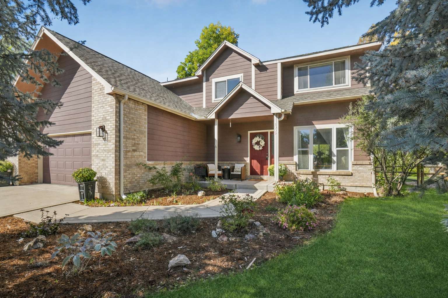 The image showcases the front exterior of a two-story home with a well-maintained lawn and landscaping. The house features a combination of brown siding and brick accents, a red front door with a wreath, and a covered entryway with seating. The overall impression is one of a welcoming and well-cared-for family home.