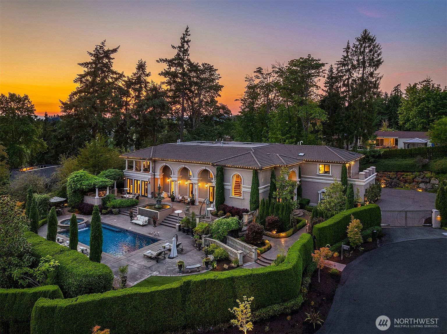 This stunning aerial view captures a luxurious Mediterranean-style estate at sunset, featuring a sprawling residence with a tiled roof and elegant arched architectural details. The property boasts a beautifully landscaped backyard complete with a sparkling swimming pool, a tiered stone fountain, and meticulously manicured hedges. The warm glow of the house lights against the vibrant orange and purple twilight sky creates an inviting and sophisticated atmosphere.