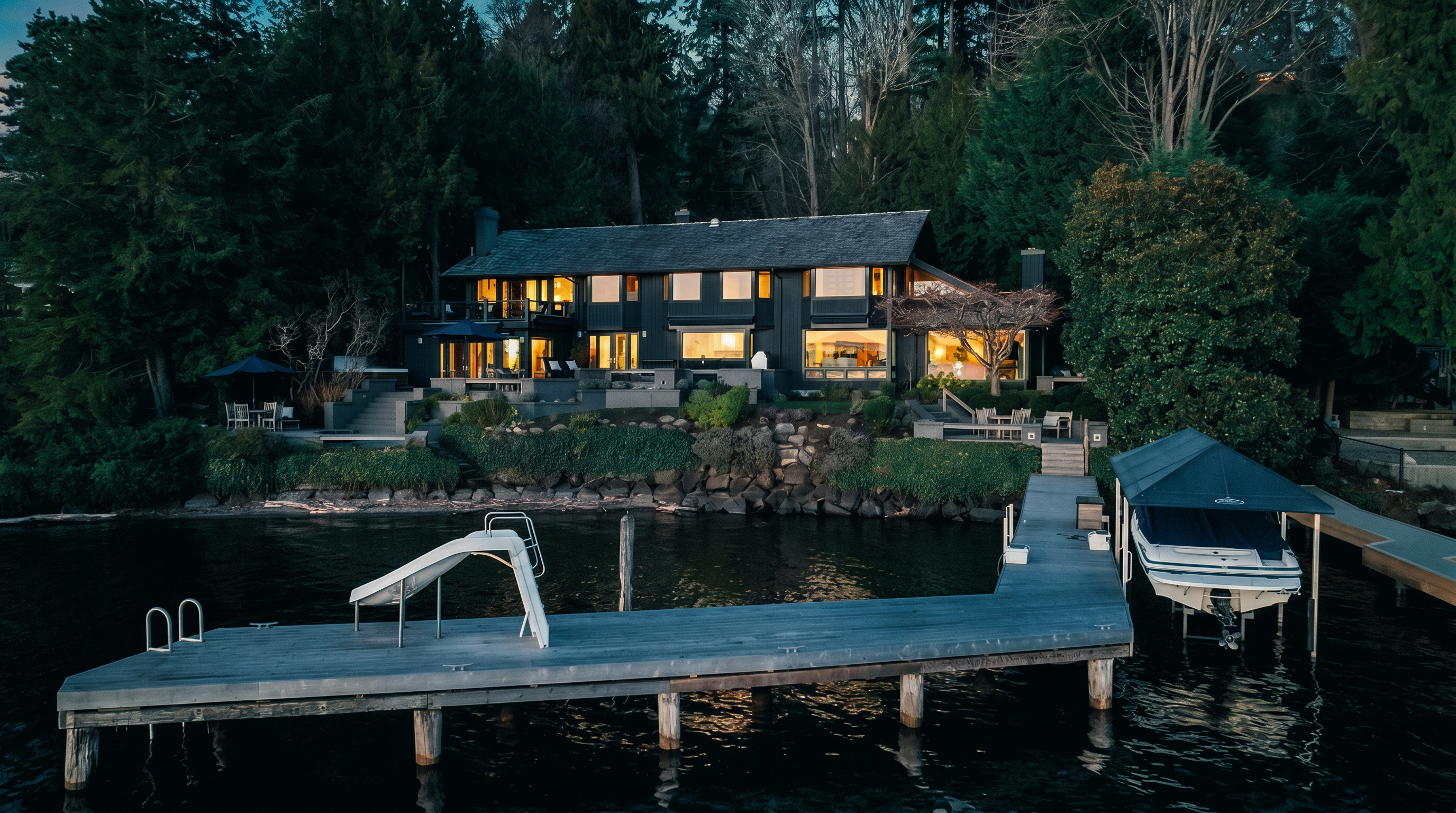 This elegant dark-toned waterfront home is captured from an elevated perspective, showcasing a sprawling multi-level deck overlooking the lake at twilight. The property features a private dock equipped with a boat lift and a water slide, seamlessly integrated into its lush, mature-forested surroundings. The warm interior lighting glowing through large windows offers a welcoming and luxurious atmosphere.