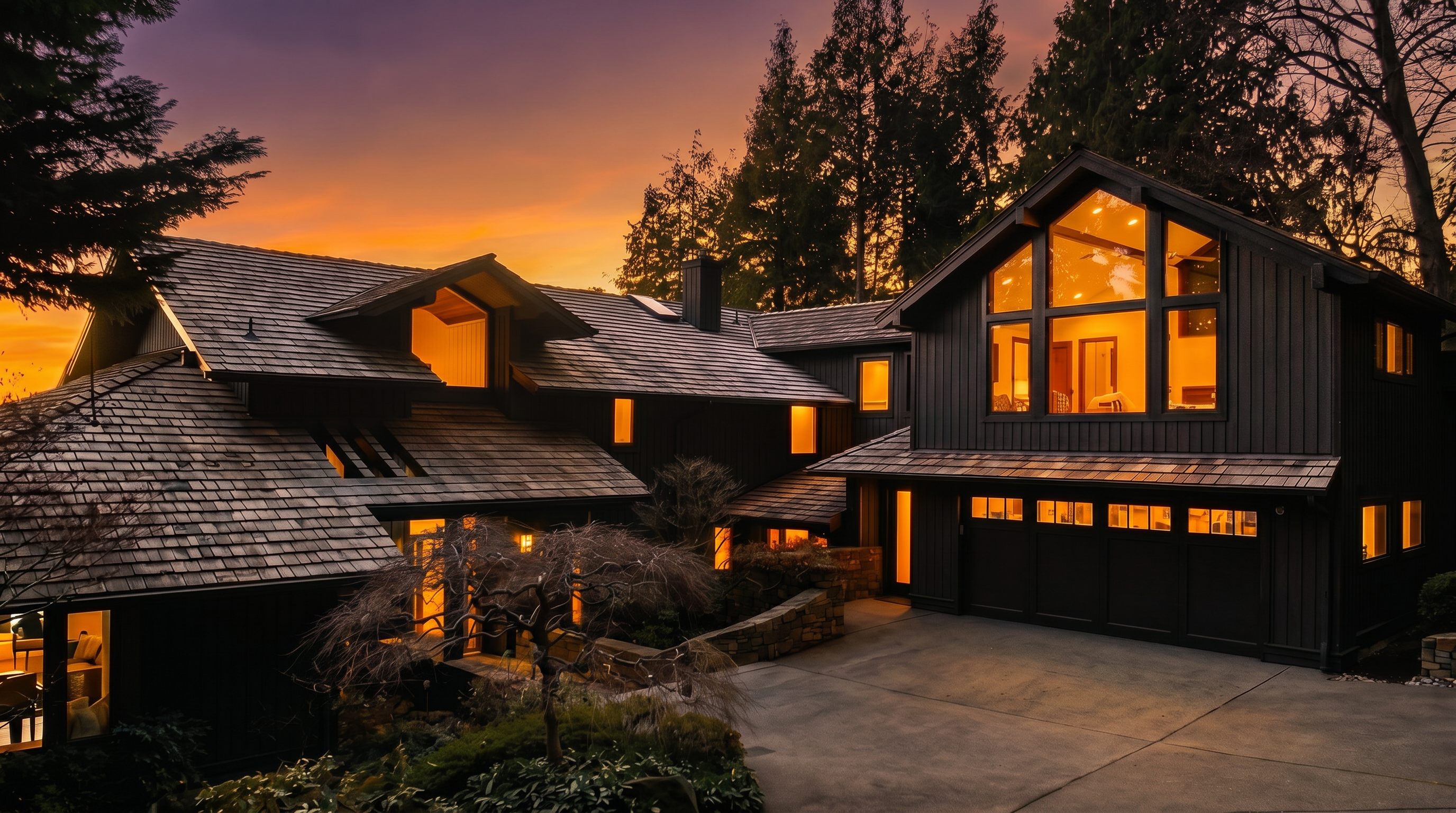 This striking modern residence features a dark-painted vertical siding aesthetic contrasted by large geometric windows that glow with warm lighting against a vibrant sunset. The architectural design includes an expansive shingled roofline, an integrated multi-car garage, and thoughtfully layered landscaping that frames the approach. The combination of its dramatic evening lighting and sophisticated, sharp-lined structure gives the property a luxurious and inviting curb appeal.
