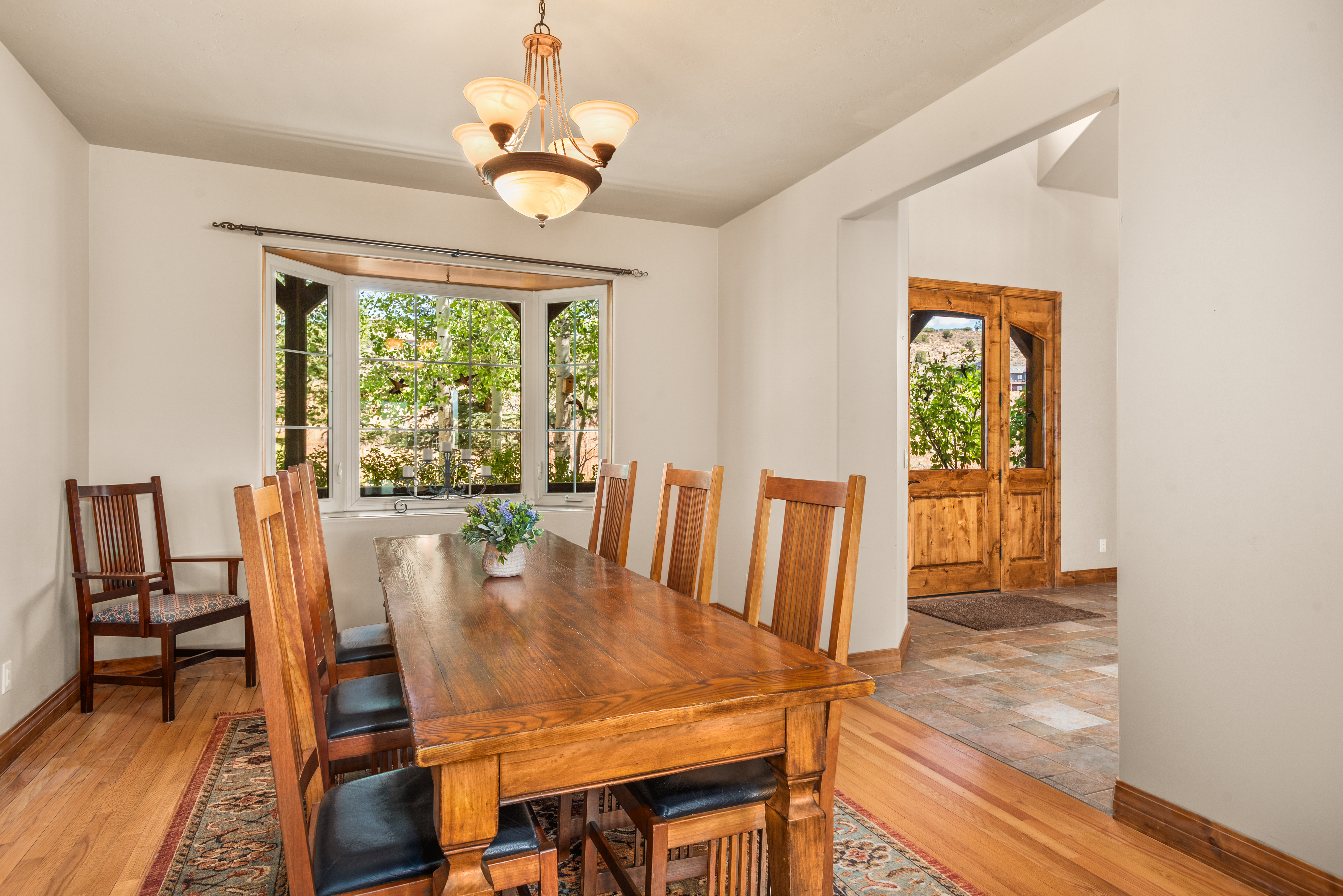 This warm and inviting dining room features a large, rustic wooden table paired with matching chairs, elegantly placed on an intricate area rug. A classic chandelier hangs above while a large bay window fills the space with natural light and views of the surrounding trees. The wood flooring and seamless transition to the tiled entryway provide a cohesive and traditional aesthetic.