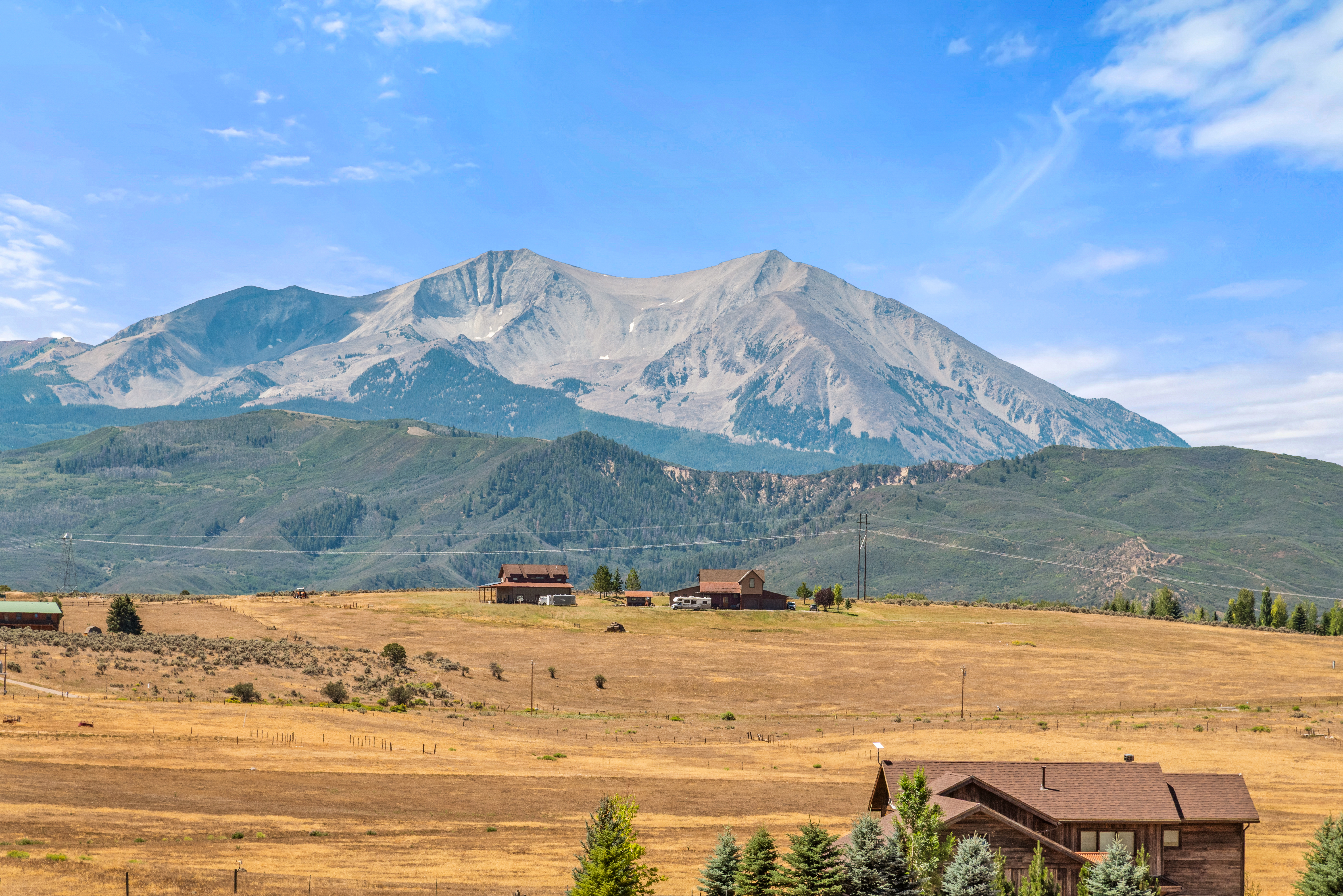 This scenic landscape captures a vast, golden-toned field with several rustic residential structures nestled against a backdrop of majestic, snow-capped mountains. The perspective offers a tranquil, expansive view typical of a rural property setting, emphasizing the property's integration with the natural topography. The clear blue sky and rolling hills enhance the picturesque, quiet appeal of this countryside estate.