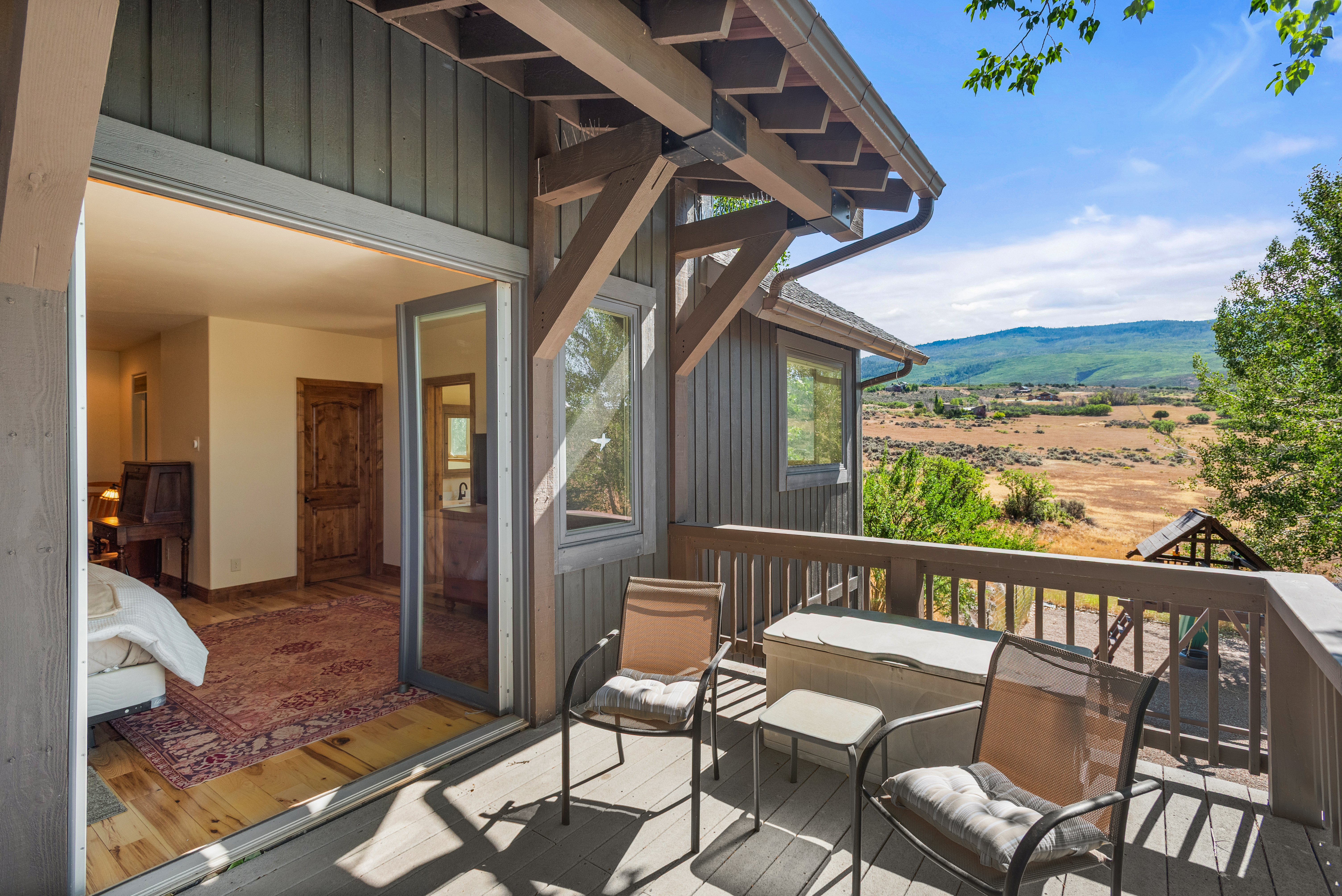 This image features a rustic wooden deck overlooking a scenic landscape with rolling hills and distant mountains. The deck is furnished with two chairs and a small table, and provides seamless indoor-outdoor access to a bedroom through a large sliding glass door. The charming architectural details, such as the exposed timber roof supports, enhance the home's mountain-style aesthetic.