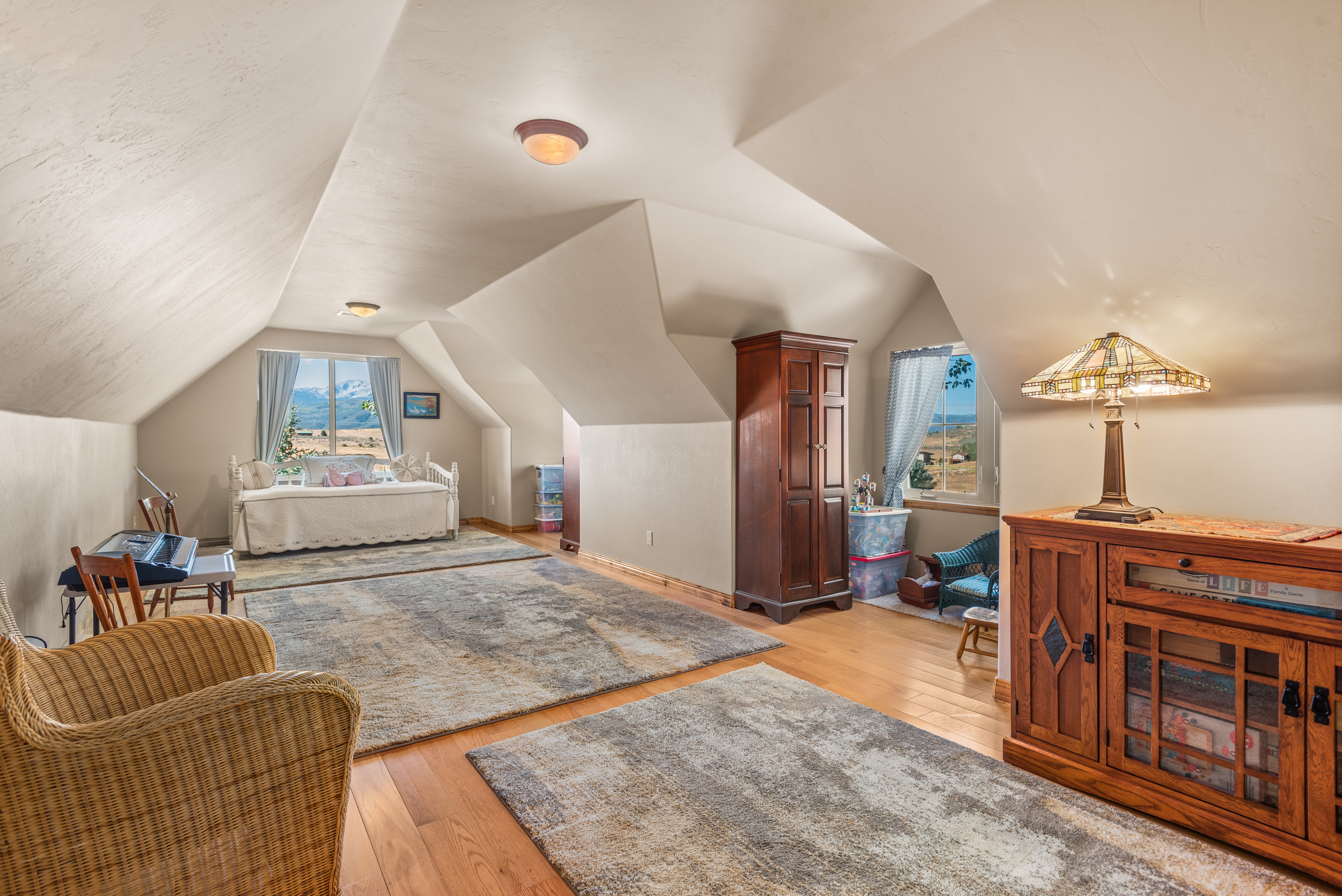 This spacious and sunlit attic bedroom features high, vaulted ceilings and warm wood flooring, creating an inviting and airy atmosphere. The room is thoughtfully furnished with a cozy daybed, a traditional wooden cabinet, and a Tiffany-style lamp, making it a versatile space perfect for a sleeping area or a serene lounge. With multiple windows offering beautiful scenic views, the room feels connected to the outdoors while maintaining a comfortable, secluded retreat feel.
