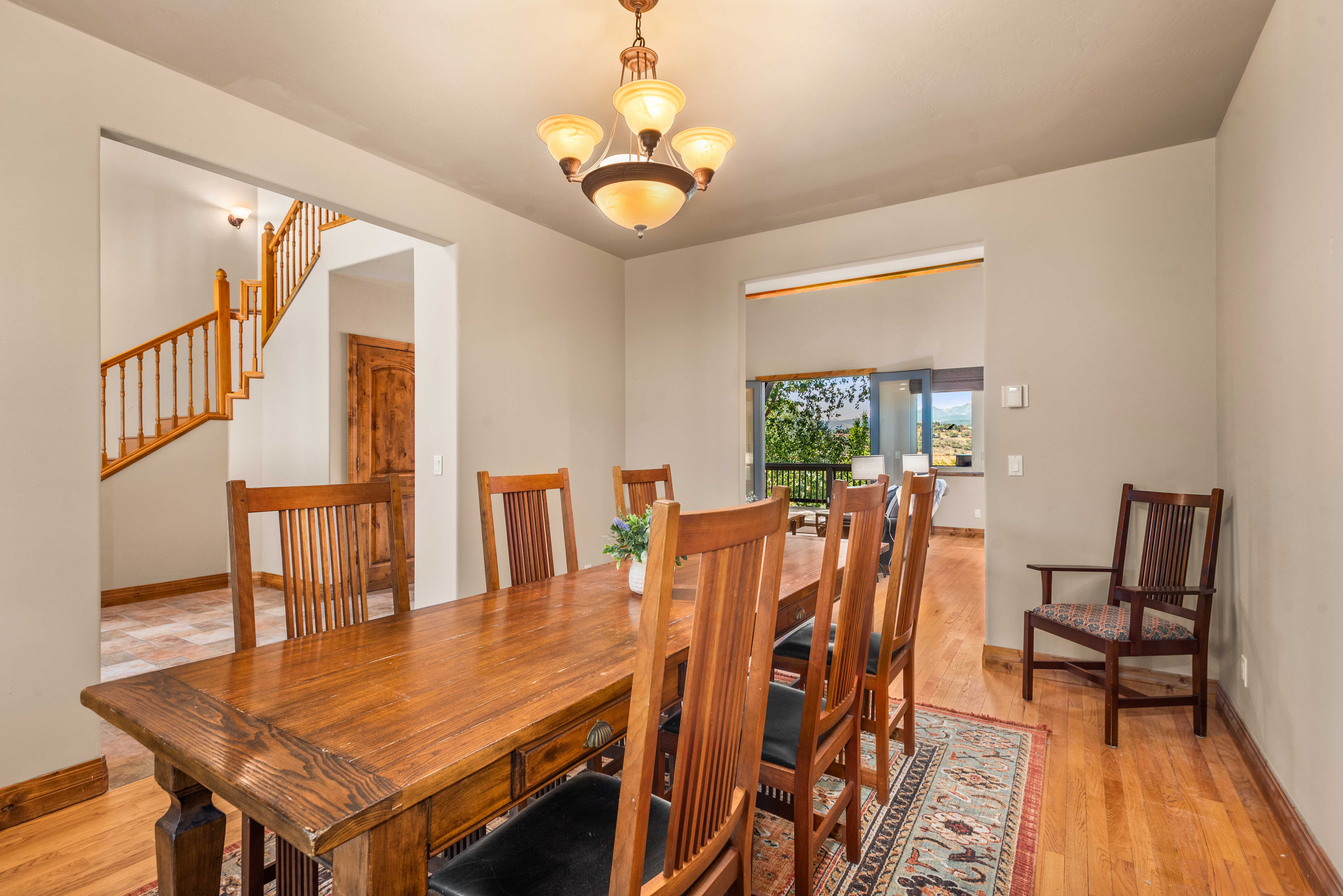 This spacious dining room features a warm, traditional aesthetic centered around a large solid wood table with matching slatted-back chairs. The room is anchored by a patterned area rug and illuminated by an elegant multi-light chandelier, while a glimpse of the staircase and an outdoor deck adds visual depth. The open floor plan and natural wood tones create a welcoming and comfortable atmosphere for gatherings.