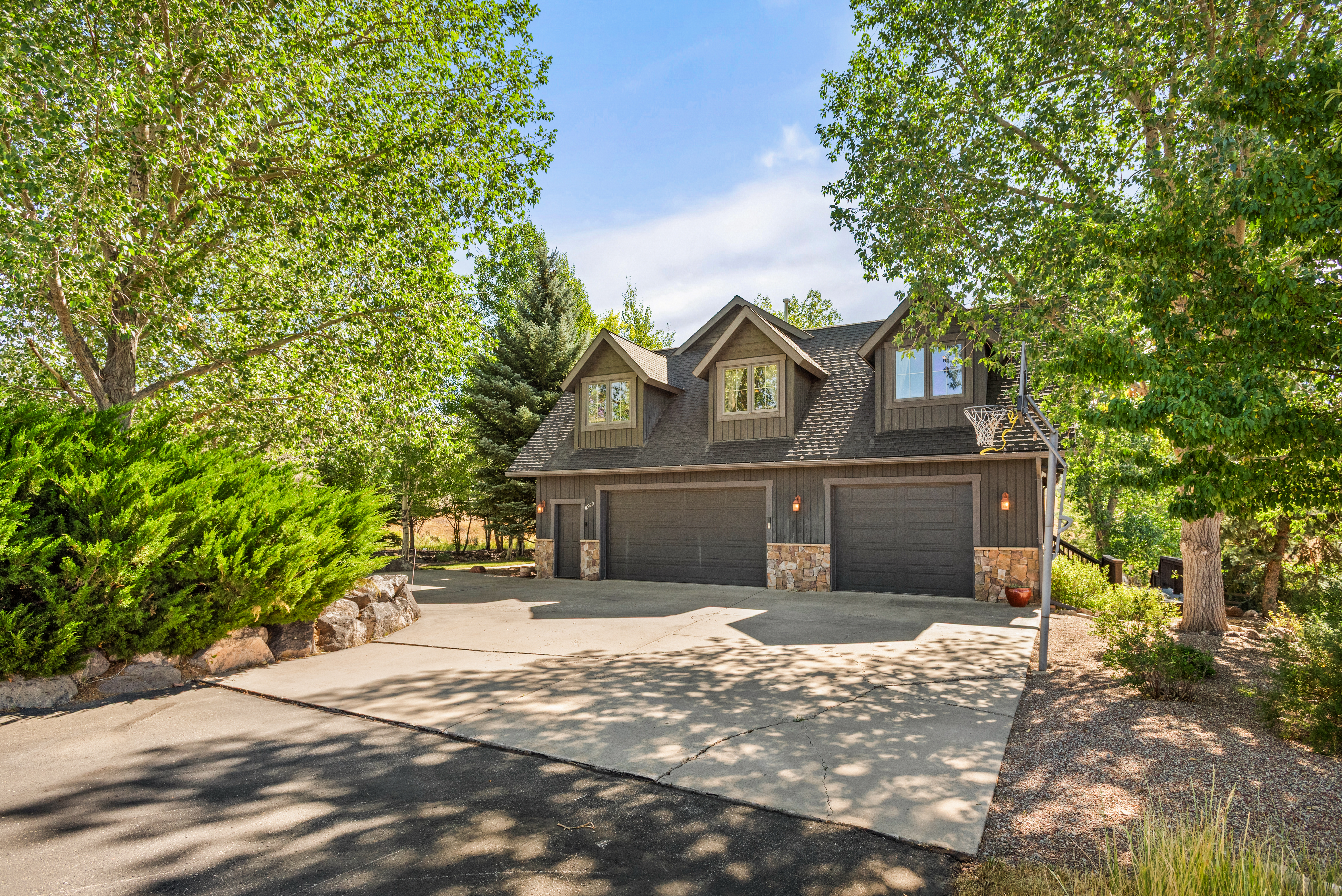 This image displays the exterior of a charming two-story detached garage featuring wood siding, stone accents, and three gabled dormers above the roofline. The structure includes two spacious garage doors, a pedestrian side entrance, and a concrete driveway complemented by mature trees and landscaped rocks. The overall property has a rustic, mountain-retreat aesthetic well-suited for additional vehicle storage or shop space.