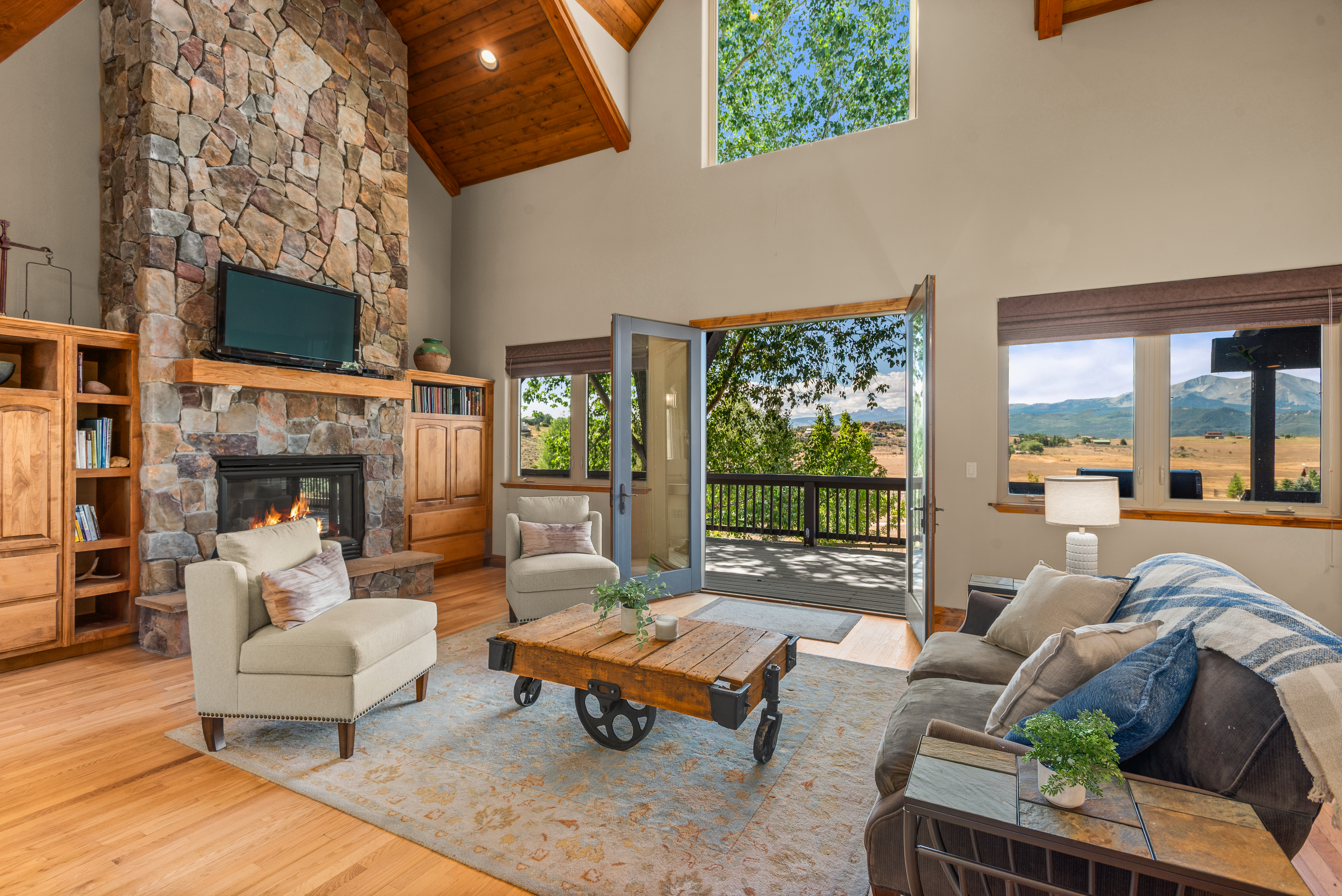 This inviting living room features a striking floor-to-ceiling stone fireplace as its focal point, complemented by vaulted wood-paneled ceilings. The space is naturally brightened by expansive windows and double doors that open onto a deck, offering sweeping views of the surrounding mountainous landscape. The rustic-cozy aesthetic is enhanced by hardwood floors, a vintage industrial-style coffee table, and comfortable, neutral-toned furnishings.