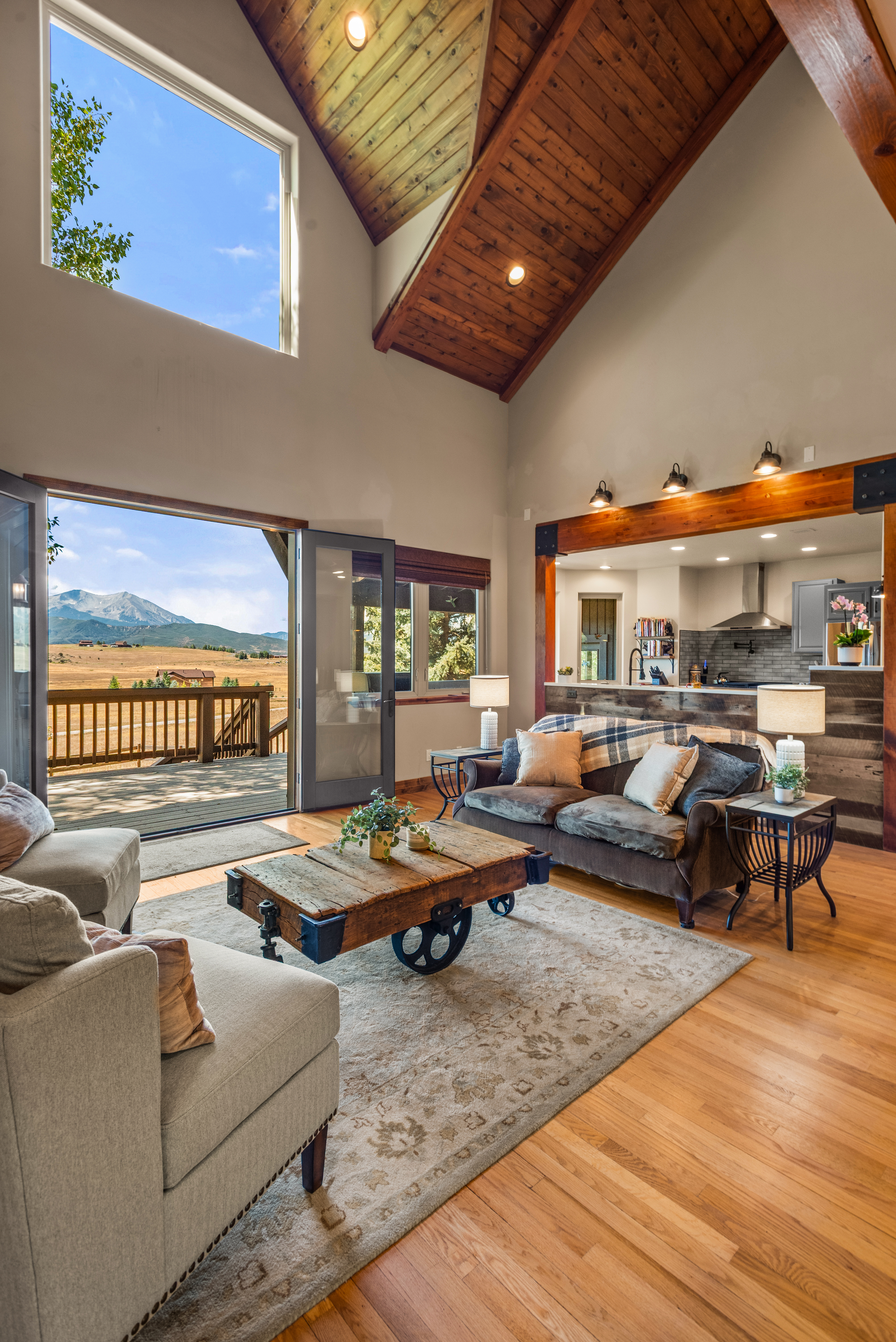 This inviting living room features vaulted wooden ceilings, hardwood flooring, and a unique industrial-style coffee table with wheels. An expansive open-door concept leads to a spacious deck with stunning mountain views, seamlessly blending interior comfort with outdoor scenery. A separate, cozy kitchen space is visible in the background, making this an ideal layout for entertaining and mountain living.