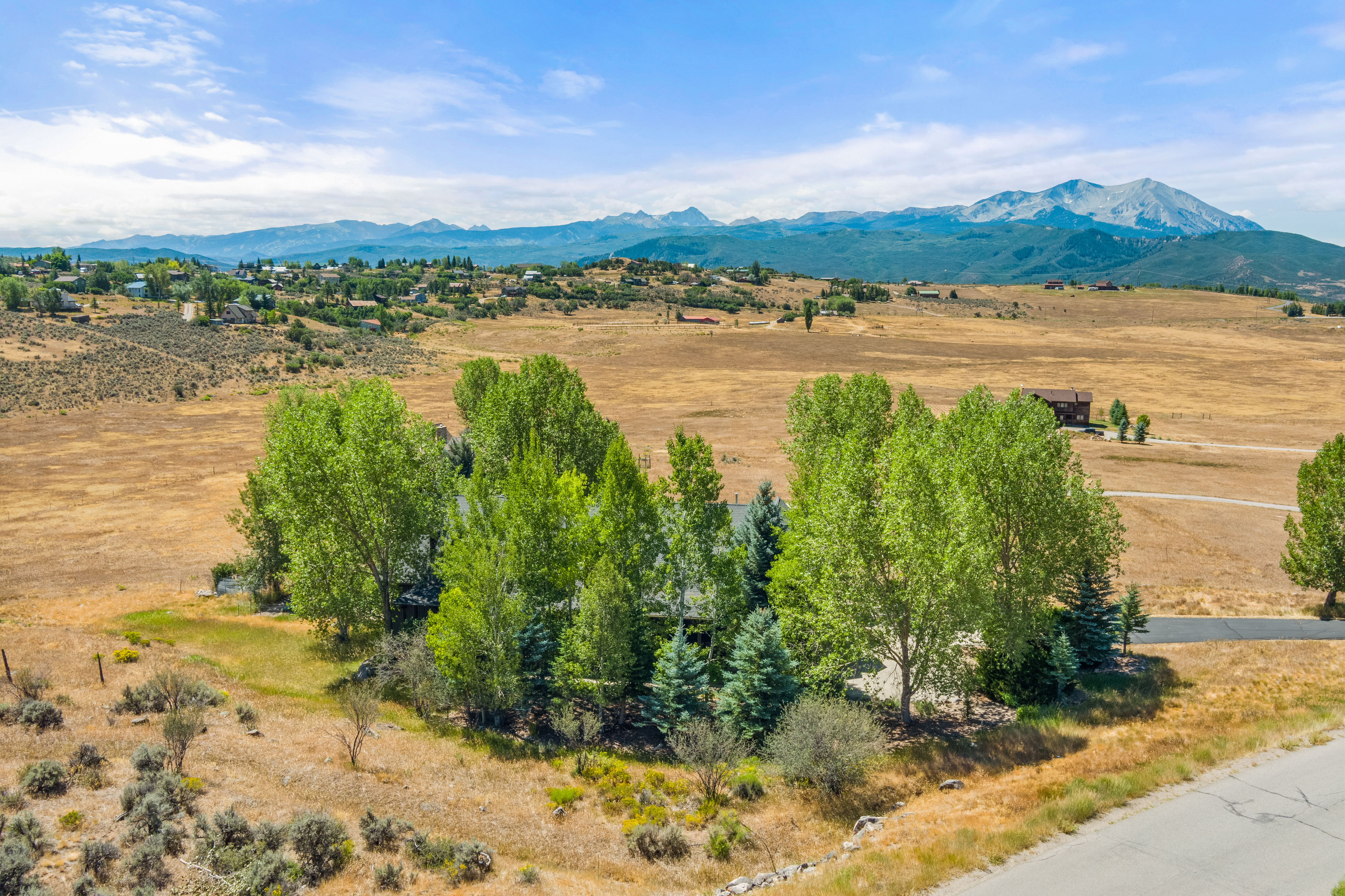 This stunning wide-angle aerial shot showcases a secluded home nestled within a dense cluster of lush green trees, providing privacy and shade. The vast, sprawling landscape features a dry, golden prairie setting with majestic mountain peaks towering in the background under a expansive blue sky. The property offers a serene, rustic retreat atmosphere, perfectly blending into its natural, mountainous environment.