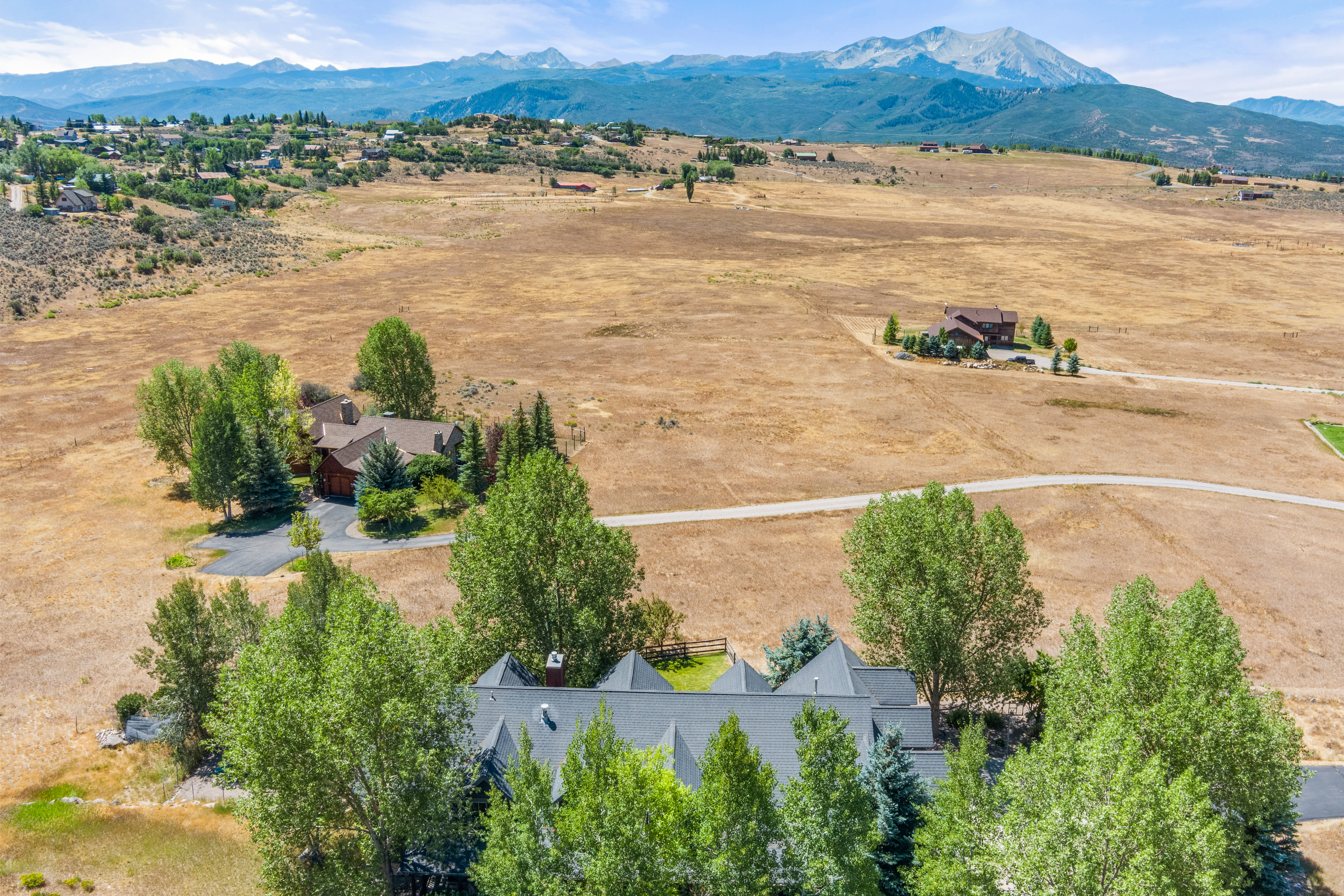 This high-angle, aerial perspective showcases a sprawling property featuring two prominent detached houses set amidst a vast, dry, golden-grass meadow. The expansive landscape is framed by a stunning backdrop of rugged mountain ranges under a clear, bright sky. The property includes mature deciduous and evergreen trees surrounding the residences, connected by winding rural roads that enhance the peaceful, secluded mountain estate aesthetic.