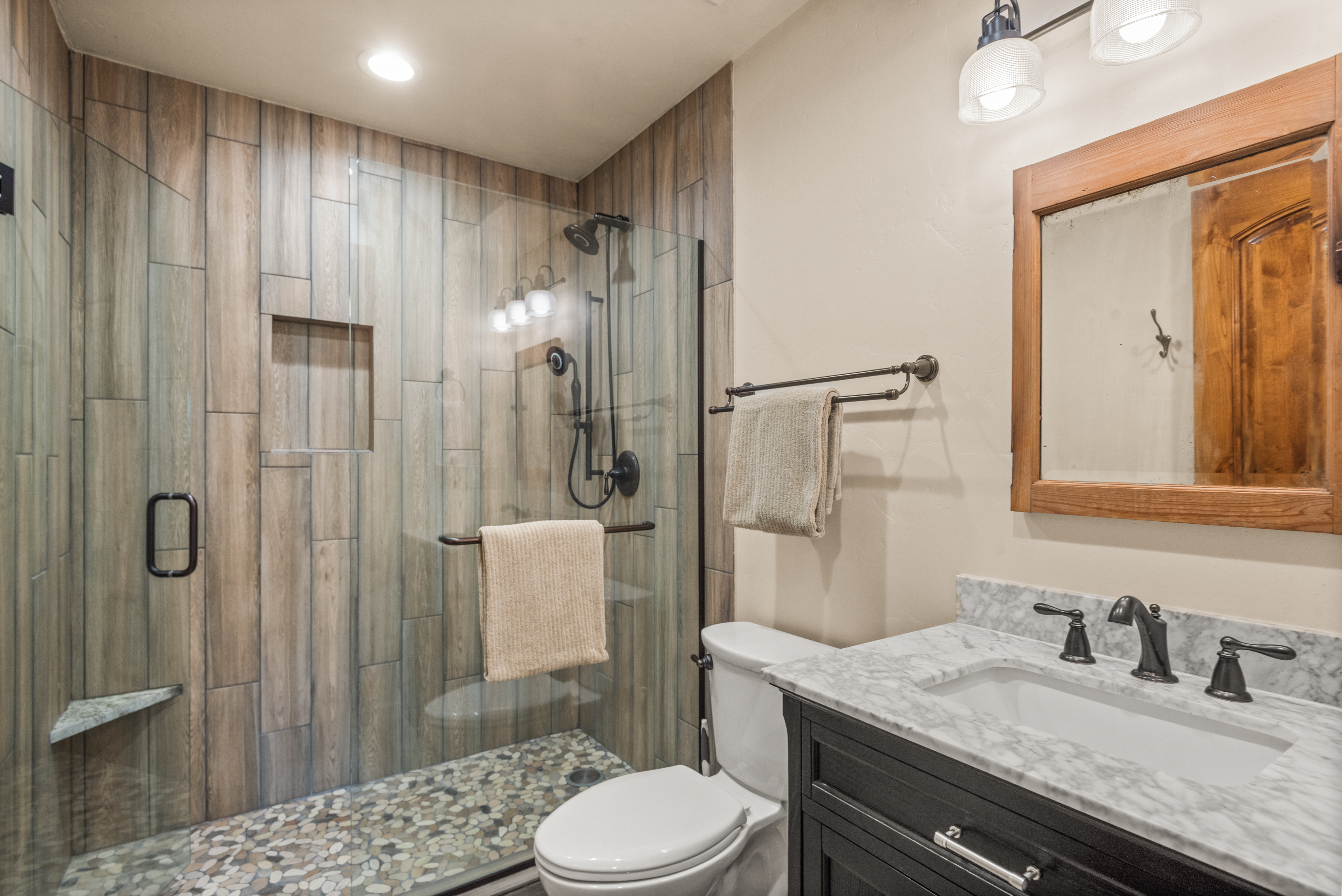 This bathroom features a contemporary design with a walk-in shower encased in glass, highlighting a warm wood-look tile surround and a pebble-stone floor. The vanity includes a stone countertop with a dark cabinet, complemented by a simple wooden-framed mirror and bronze fixtures. The overall aesthetic is rustic, modern, and inviting.