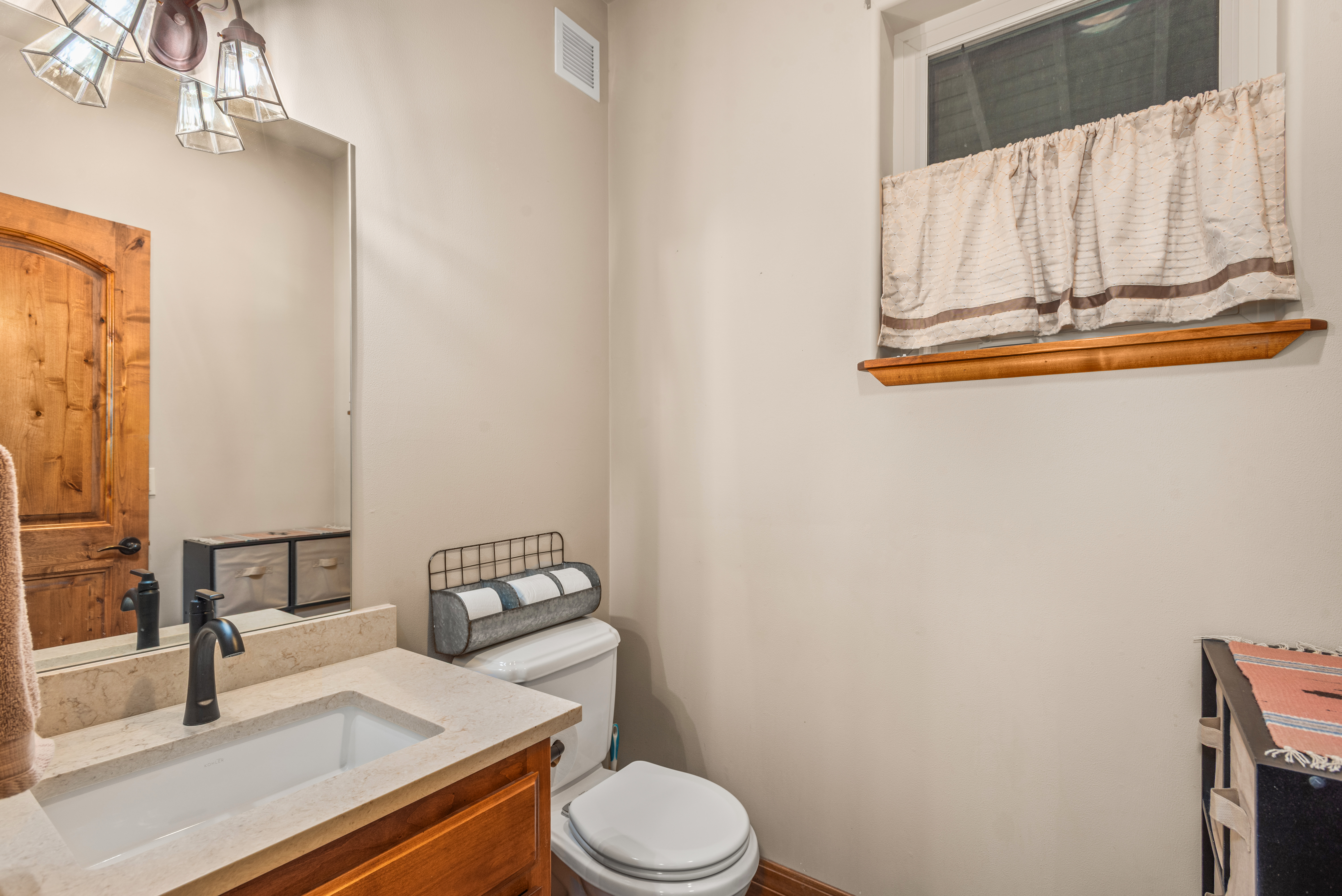 This functional bathroom features a neutral color palette with a wooden vanity, a stone-style countertop, and a modern faucet. The space includes a white toilet topped with a galvanized metal toilet paper organizer, a wall-mounted mirror, and a window with a simple valence curtain. The overall impression is a cozy and practical guest restroom with rustic-inspired accents.