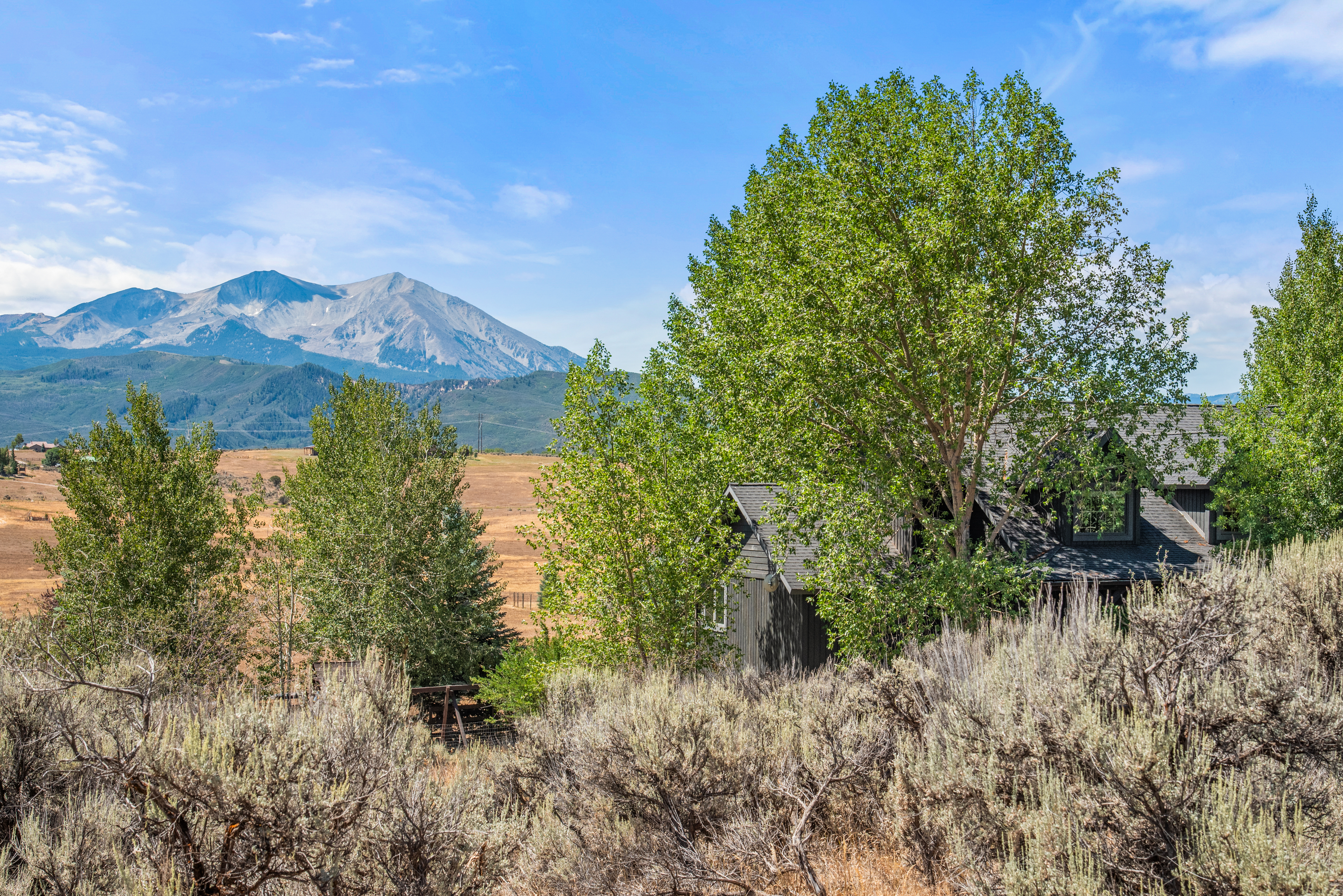This capture showcases the side profile of a dark-toned home nestled among lush, green aspen trees. The property is set against a magnificent backdrop of a prominent mountain range and expansive, dry-grass plains. The natural landscape and serene vista highlight the mountain-rustic appeal of the residence.