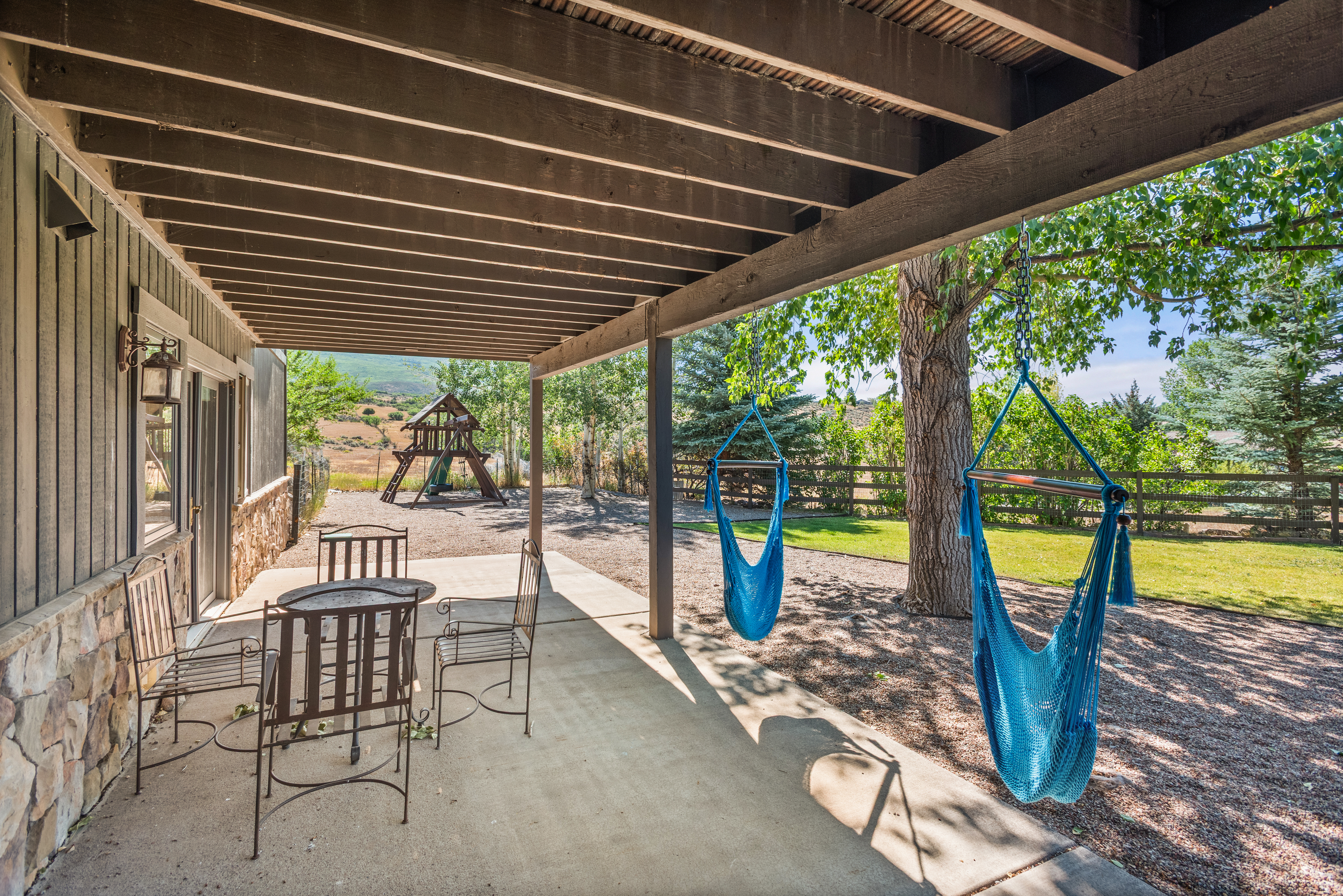 This covered backyard patio offers a relaxing space with metal tables and chairs, perfect for outdoor seating. Two blue hammock swings hang from the pergola, overlooking a spacious gravel yard with a children's play structure and mature trees. The area combines rustic stone and wooden features, creating a serene and inviting atmosphere for outdoor leisure.