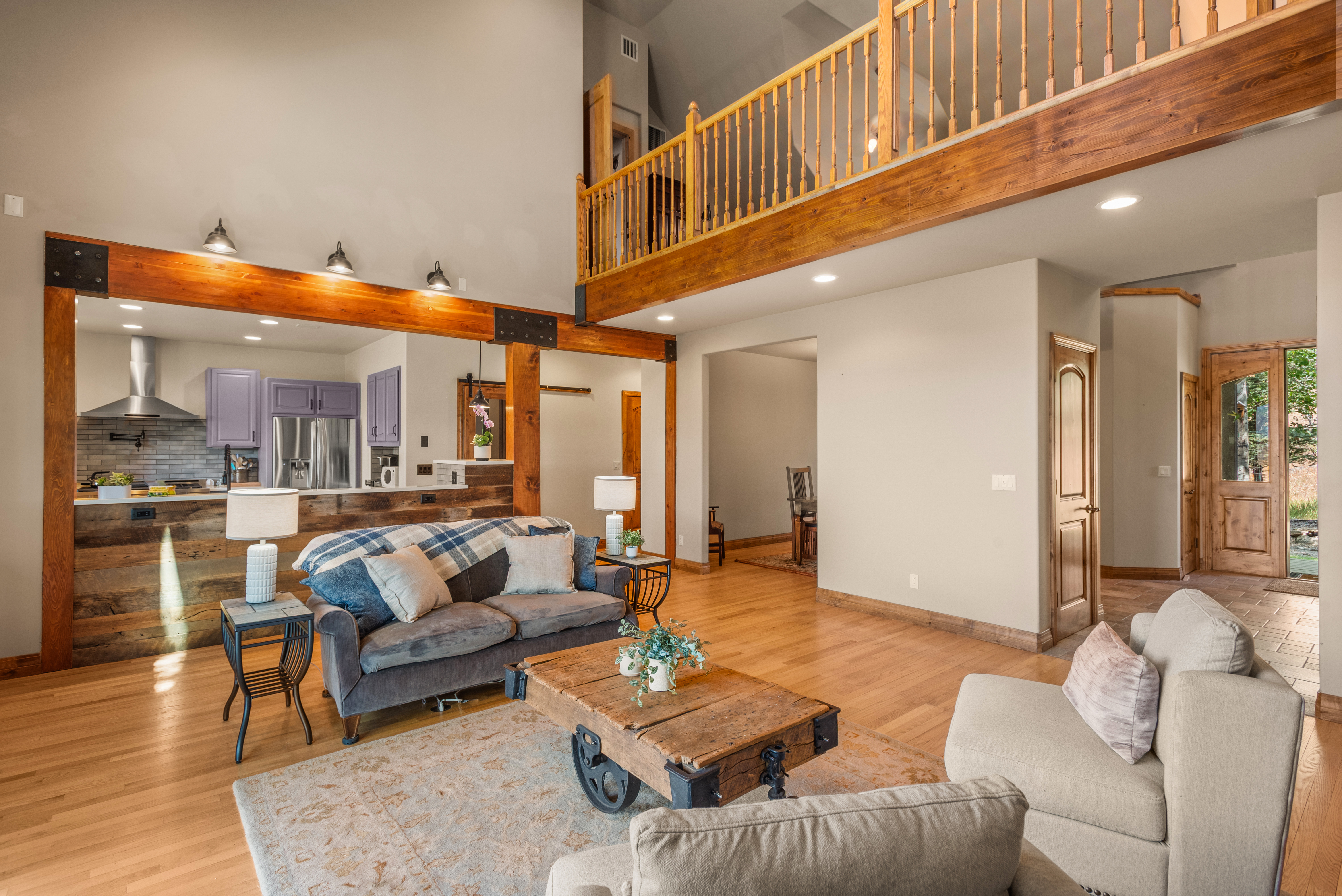 This inviting, open-concept living area features a high vaulted ceiling with a rustic wooden loft railing, perfectly blending industrial beam support with warmth. The space highlights a cozy sitting area with a unique wheeled coffee table, hardwood flooring, and a seamless flow into a kitchen with light purple cabinetry. The overall aesthetic is a harmonious mix of rustic charm and modern design, ideal for comfortable family living and entertaining.