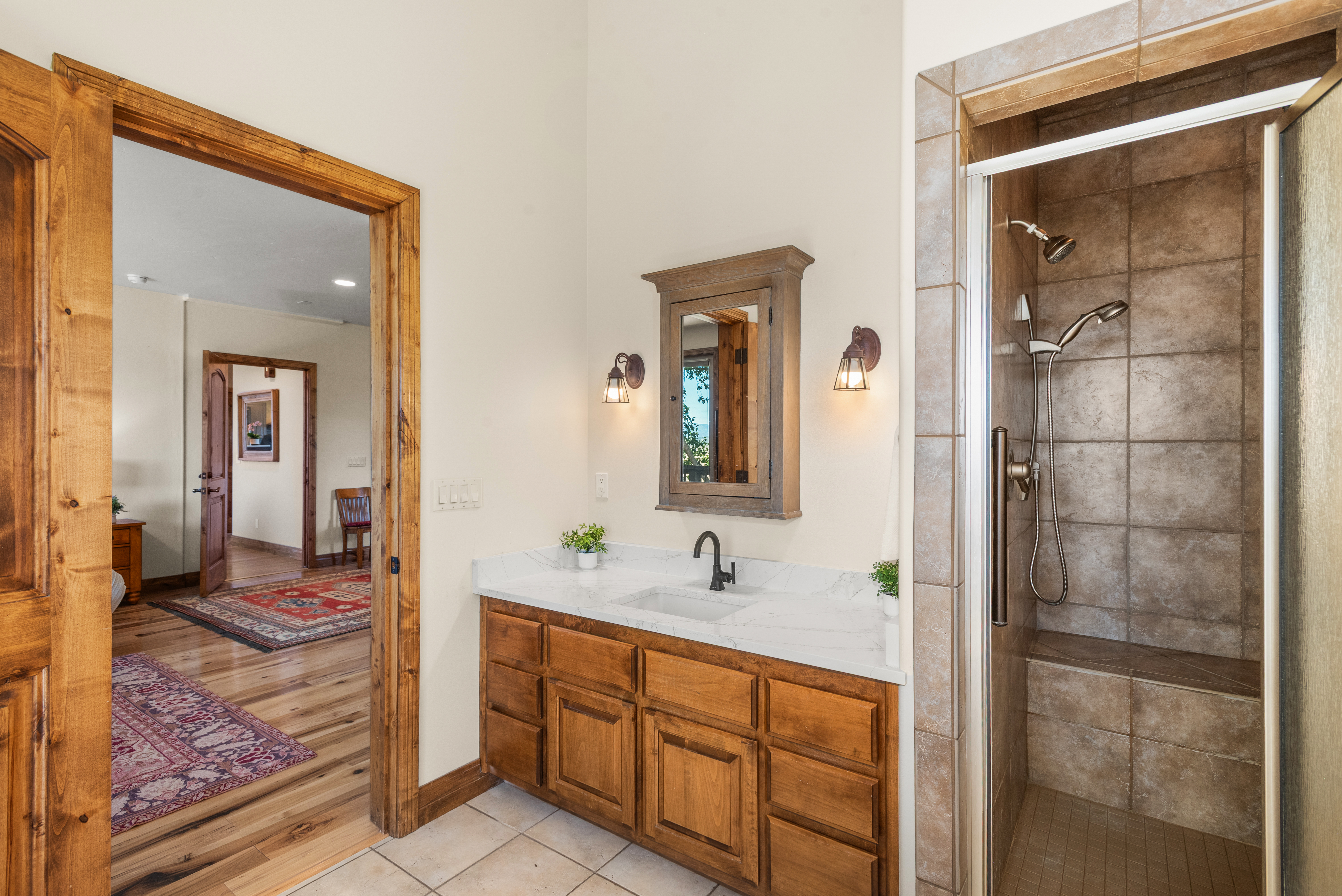 This functional bathroom features a warm, rustic aesthetic with natural wood cabinetry and a marble-look countertop. It includes a walk-in shower with tiled walls and a bench, while an open doorway provides a view into a carpeted hallway, adding to the home's open floor plan and cohesive wood-trimmed style.