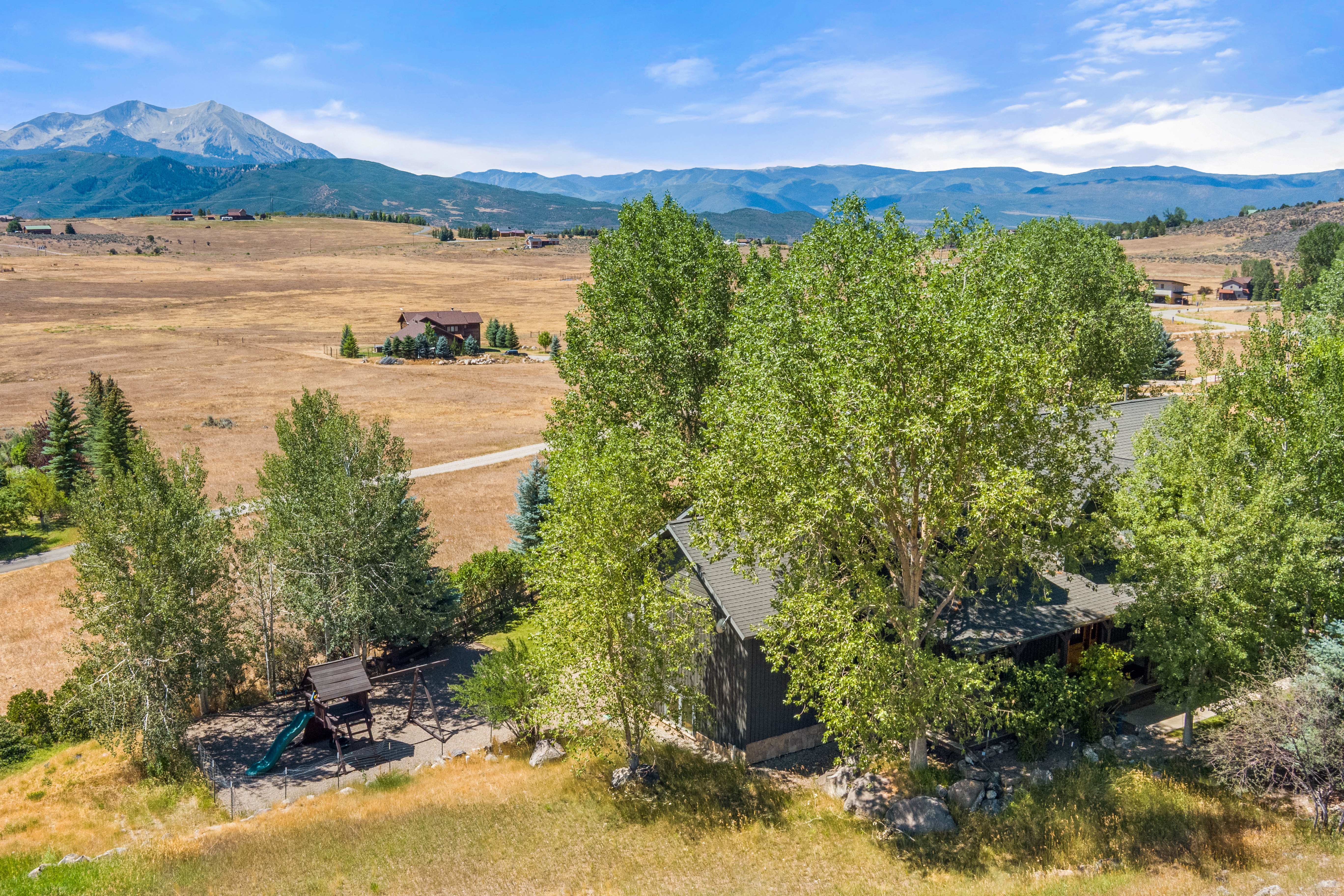 This elevated aerial view showcases a residential property nestled in a sprawling, dry mountainous landscape. The home is surrounded by mature trees, features a large side yard with a playground structure, and offers expansive views of a prominent mountain peak in the background. The rustic aesthetic and remote, scenic setting highlight its appeal as a peaceful retreat.