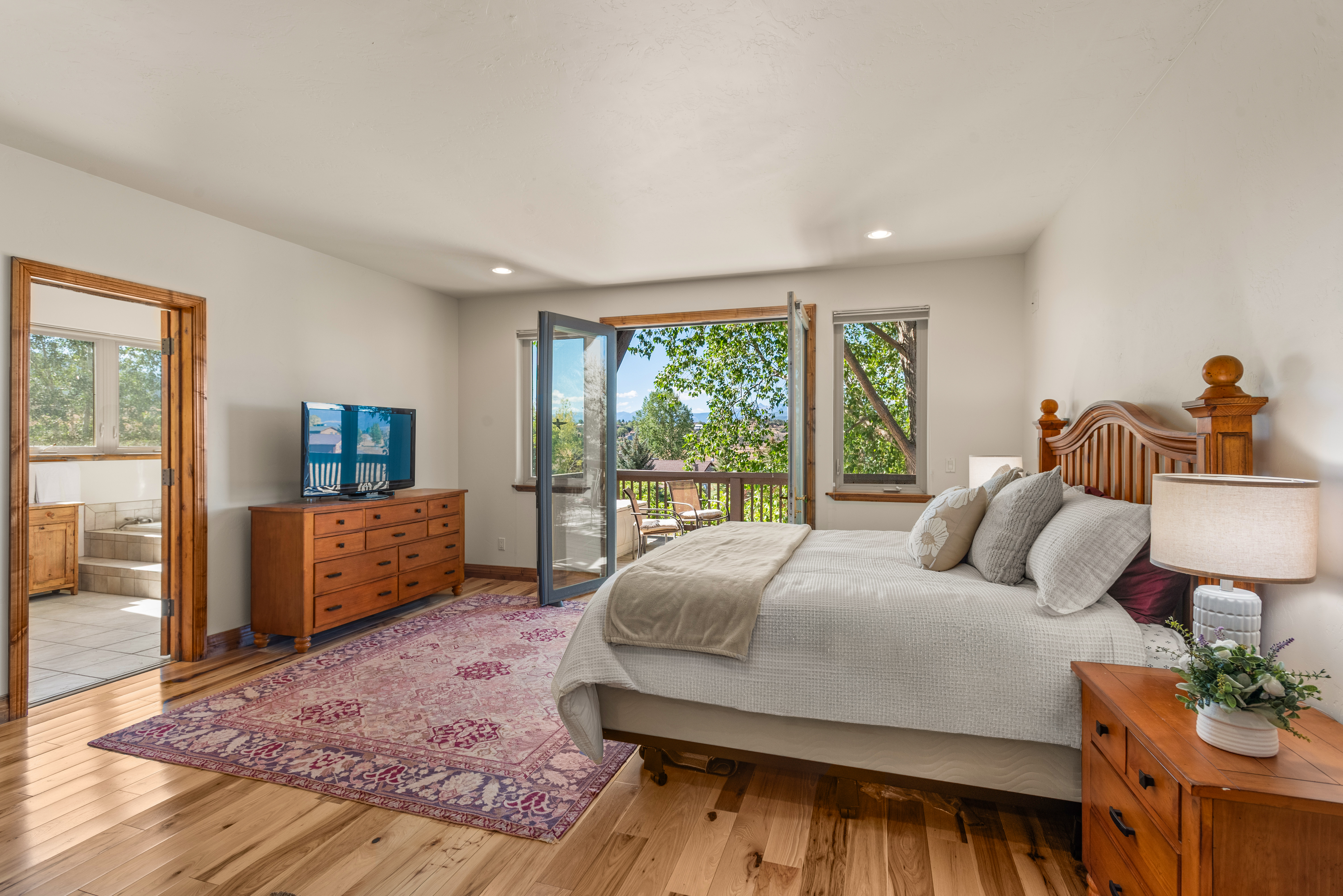This spacious primary bedroom features beautiful hardwood floors, a warm wood-framed bed, and a custom area rug, creating an inviting and comfortable atmosphere. Large glass balcony doors fill the room with natural light and offer a scenic view of the surrounding trees and outdoors. An adjoining bathroom, visible through the doorway, adds convenience to this well-appointed and serene suite.
