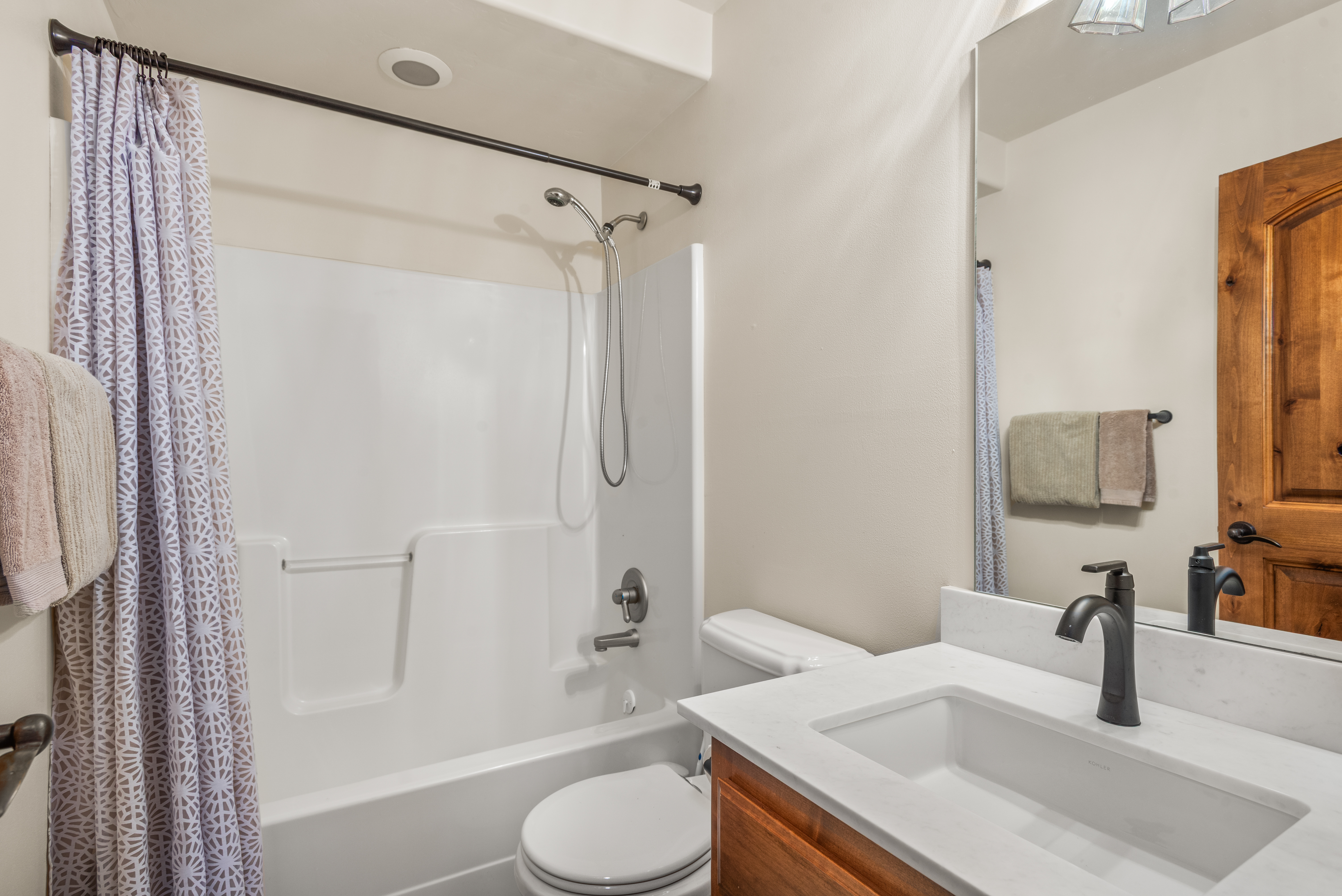 This functional bathroom features a tiled shower/tub combination with a handheld shower head and a clean, white finish. The vanity is equipped with a modern matte black faucet set against a bright, marble countertop with wooden cabinetry below. A large wall mirror and neutral wall tones create a bright and well-organized space.