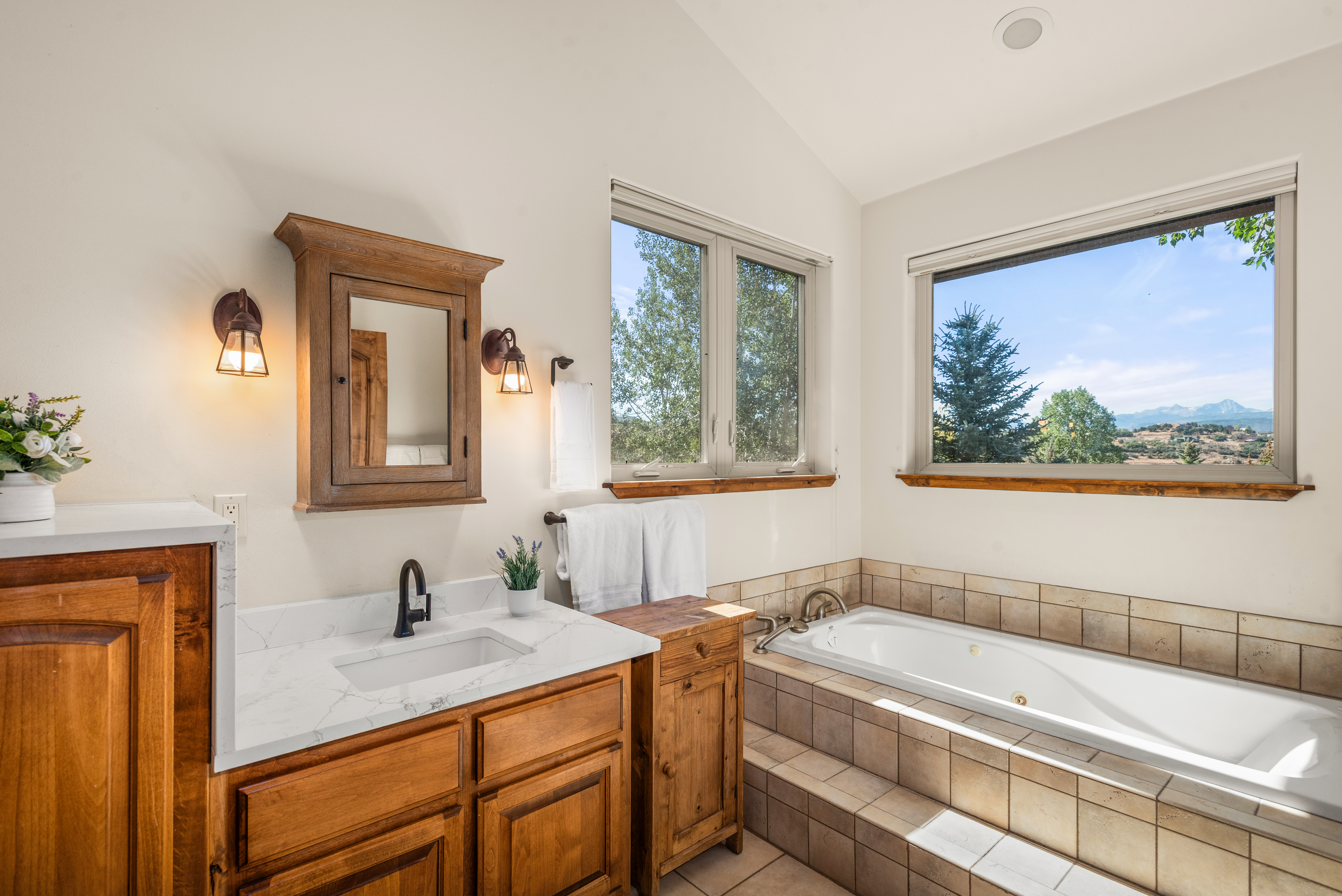 This primary bathroom features a warm, rustic aesthetic with natural wood cabinetry, white quartz countertops, and a soothing color palette. A large, tiled soaking tub is positioned perfectly to take advantage of the expansive window views of the surrounding landscape. The space is well-lit by vintage-style sconces, creating a comfortable and inviting atmosphere.