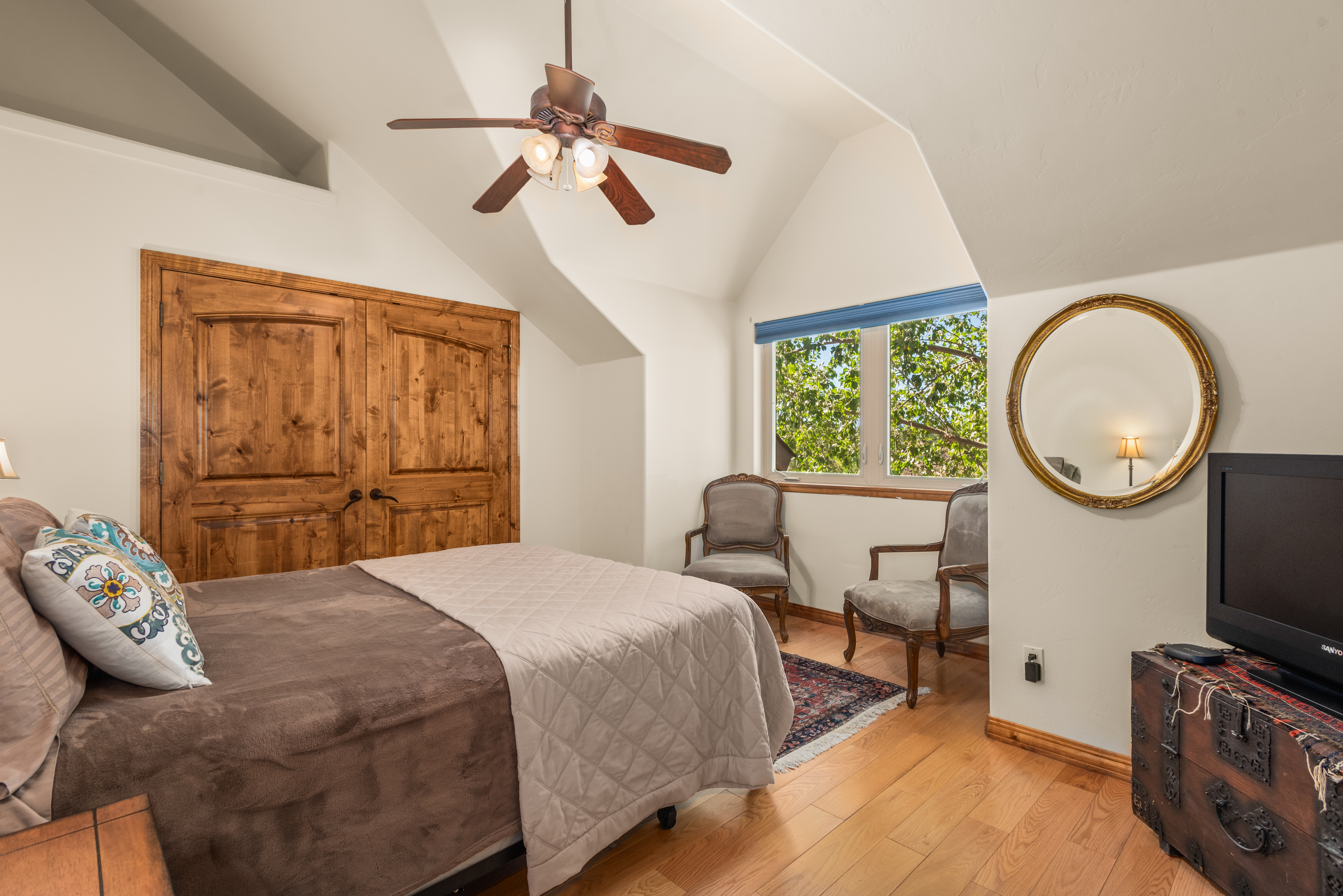 This cozy and well-appointed primary bedroom features a modern vaulted ceiling, a wooden door closet, and warm hardwood flooring. The room is accented by two vintage-style gray armchairs, an ornate gold-framed mirror, and a classic-style ceiling fan, creating a warm and inviting atmosphere. A television sits atop a decorative trunk, and the space displays a blend of rustic charm with traditional decor elements.