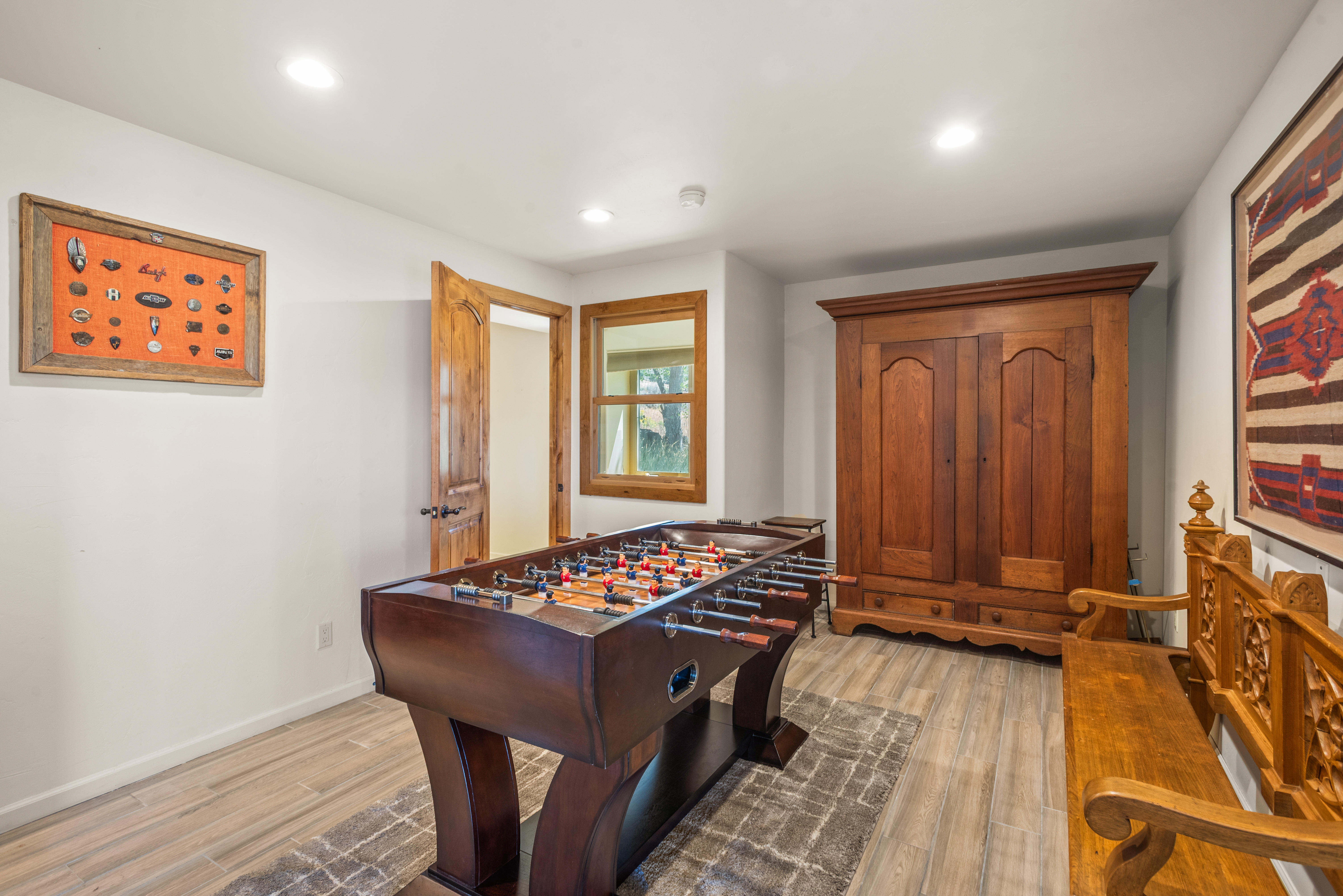 This interior space serves as a dedicated game room, featuring a prominent foosball table as the central focal point. The room includes rustic, traditional elements such as an ornate wooden bench, a large matching wardrobe, and a tapestry hanging on the wall, contrasted by modern recessed lighting and tile flooring. The space is well-lit and offers an inviting atmosphere for recreation.