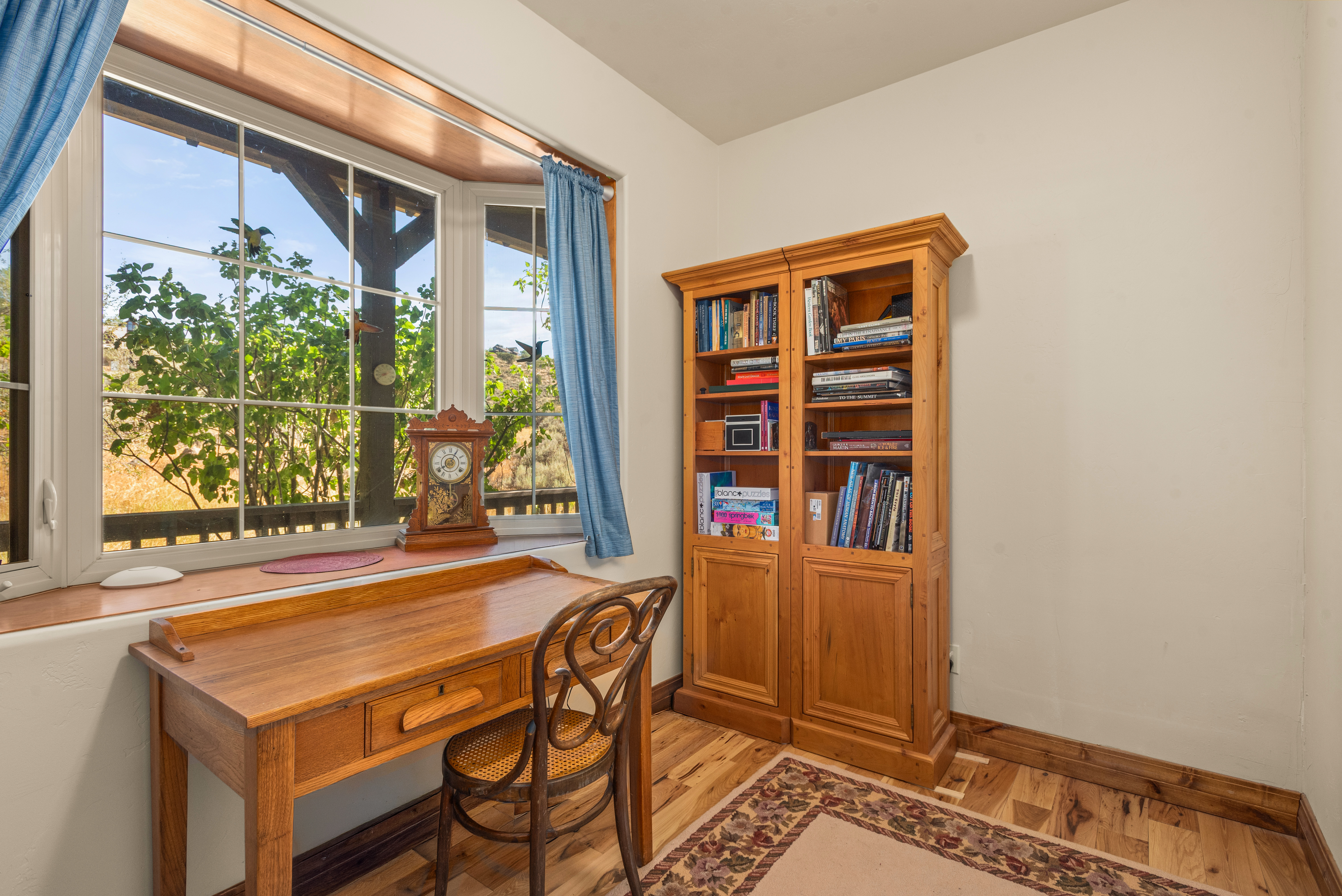 This cozy and wood-toned home office features a classic wooden desk paired with an ornate bentwood chair. A large bay window allows for ample natural light and a pleasant view, complemented by a substantial wooden bookcase filled with various volumes and puzzles. The room's warm aesthetic is rooted in its natural wood finishes and neutral walls, creating an inviting space for productivity.