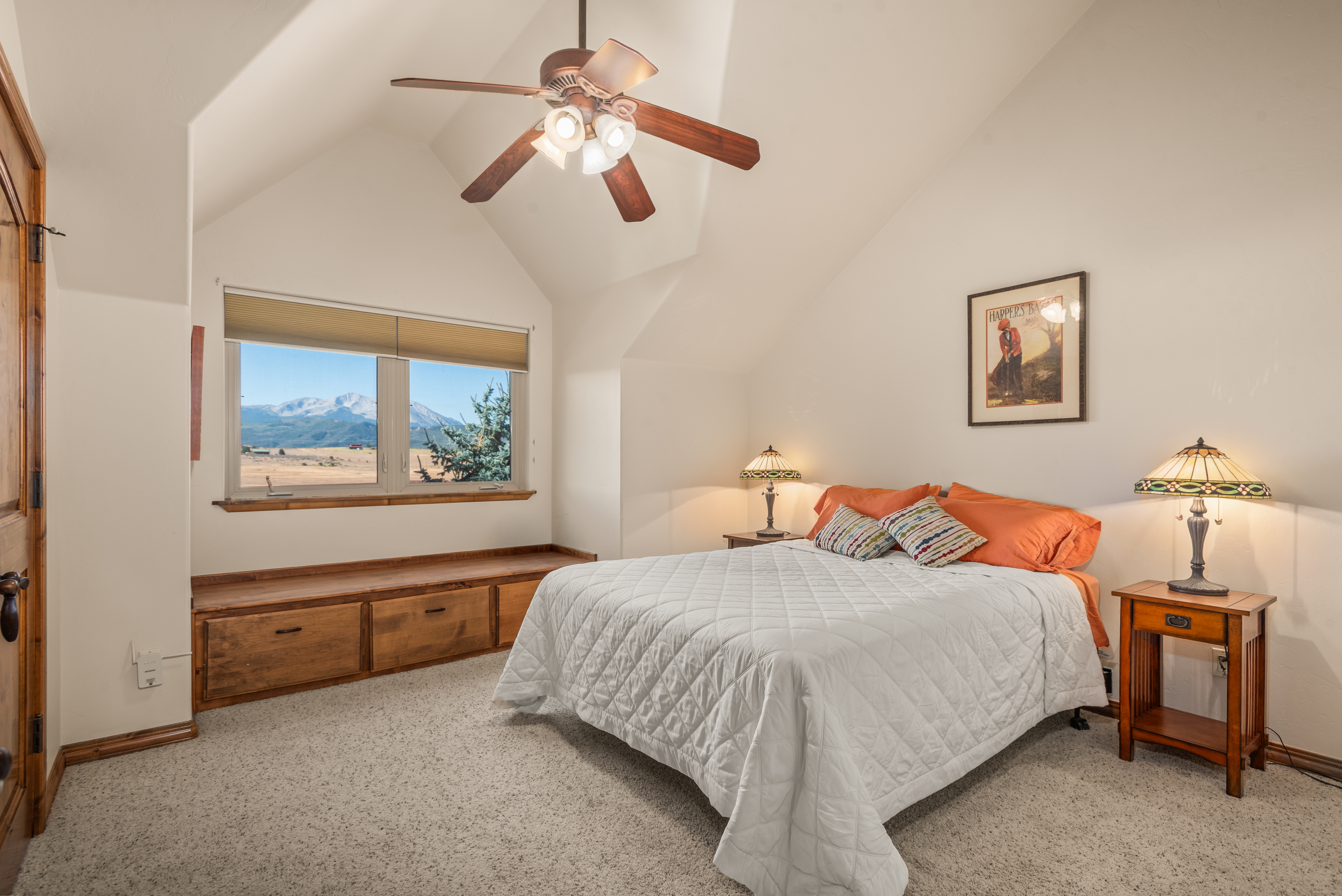 This cozy bedroom features a high, vaulted ceiling and a built-in wooden storage bench beneath a large window that offers picturesque mountain views. The room is appointment with a white quilted bedspread, orange pillows, and two stained-glass lamps, creating a warm and inviting atmosphere. The neutral carpet and clean wall lines enhance the spacious feel of this comfortable guest room.