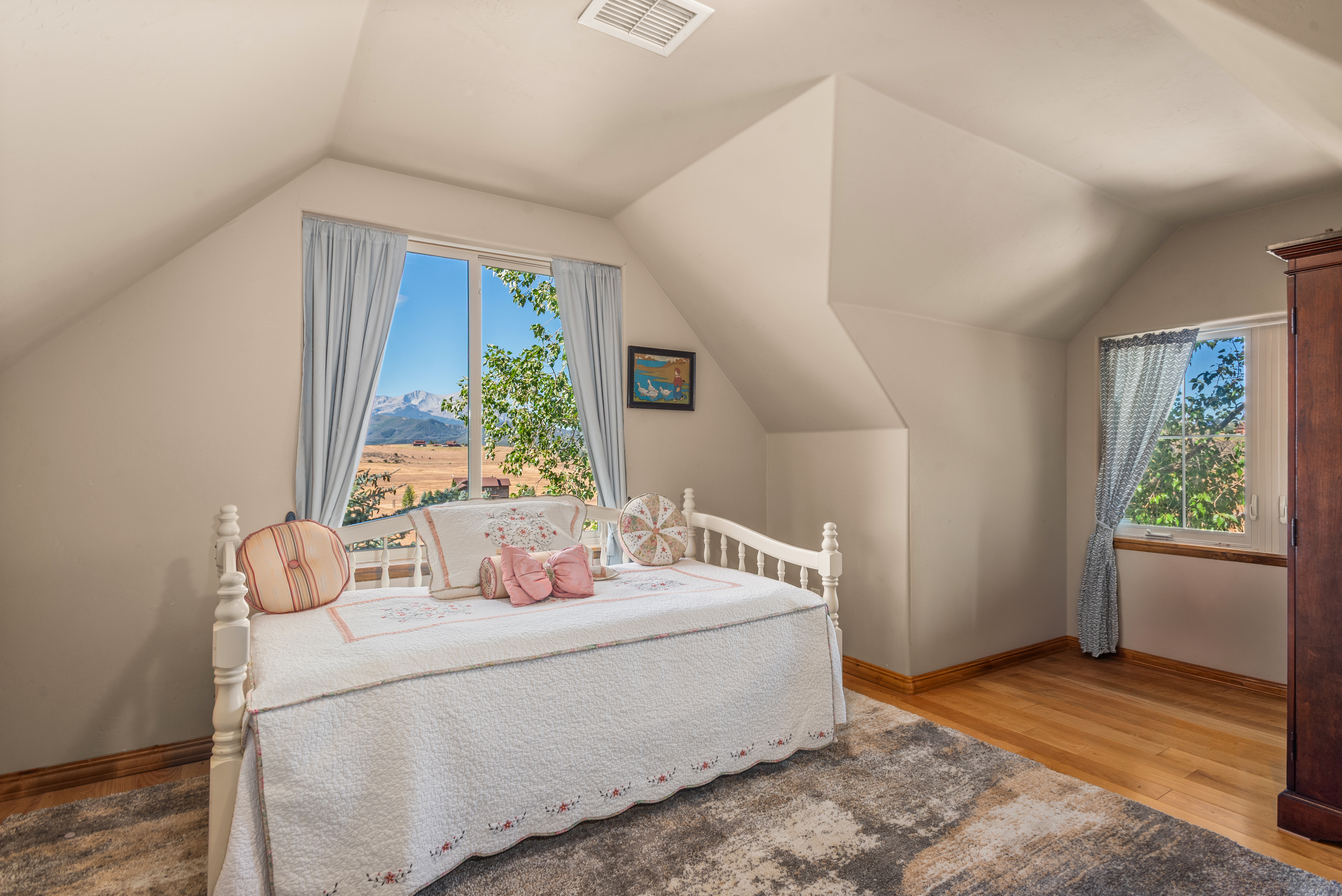 This charming guest bedroom features a white daybed with a delicate quilted coverlet and decorative pillows, set against light-colored walls and warm hardwood floors. A unique sloped ceiling adds character to the space while large windows provide scenic views of the surrounding landscape and abundant natural light. The room is finished with a neutral patterned area rug and a traditional wardrobe, creating a cozy and inviting retreat.
