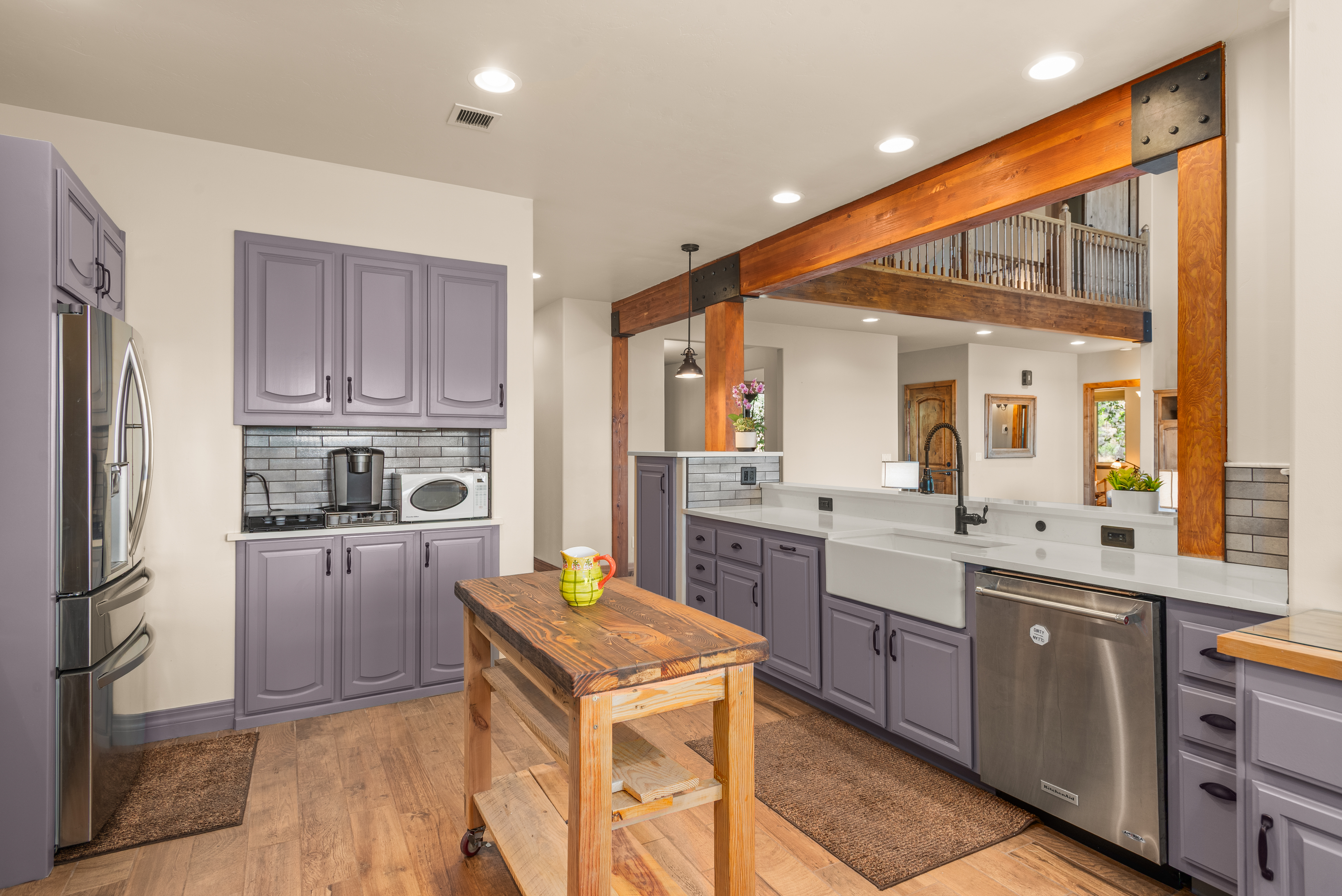 This kitchen showcases a unique rustic-modern design, featuring distinctive mauve-colored cabinetry paired with dark hardwood floors and massive wooden structural beams. The layout includes a portable wooden island, an apron-front farmhouse sink, and a coffee station, complemented by a mezzanine overlooking the kitchen that creates an open, dramatic feel. The overall aesthetic combines warm wood textures with polished stainless steel appliances and crisp white countertops, making it an inviting focal point of the home.