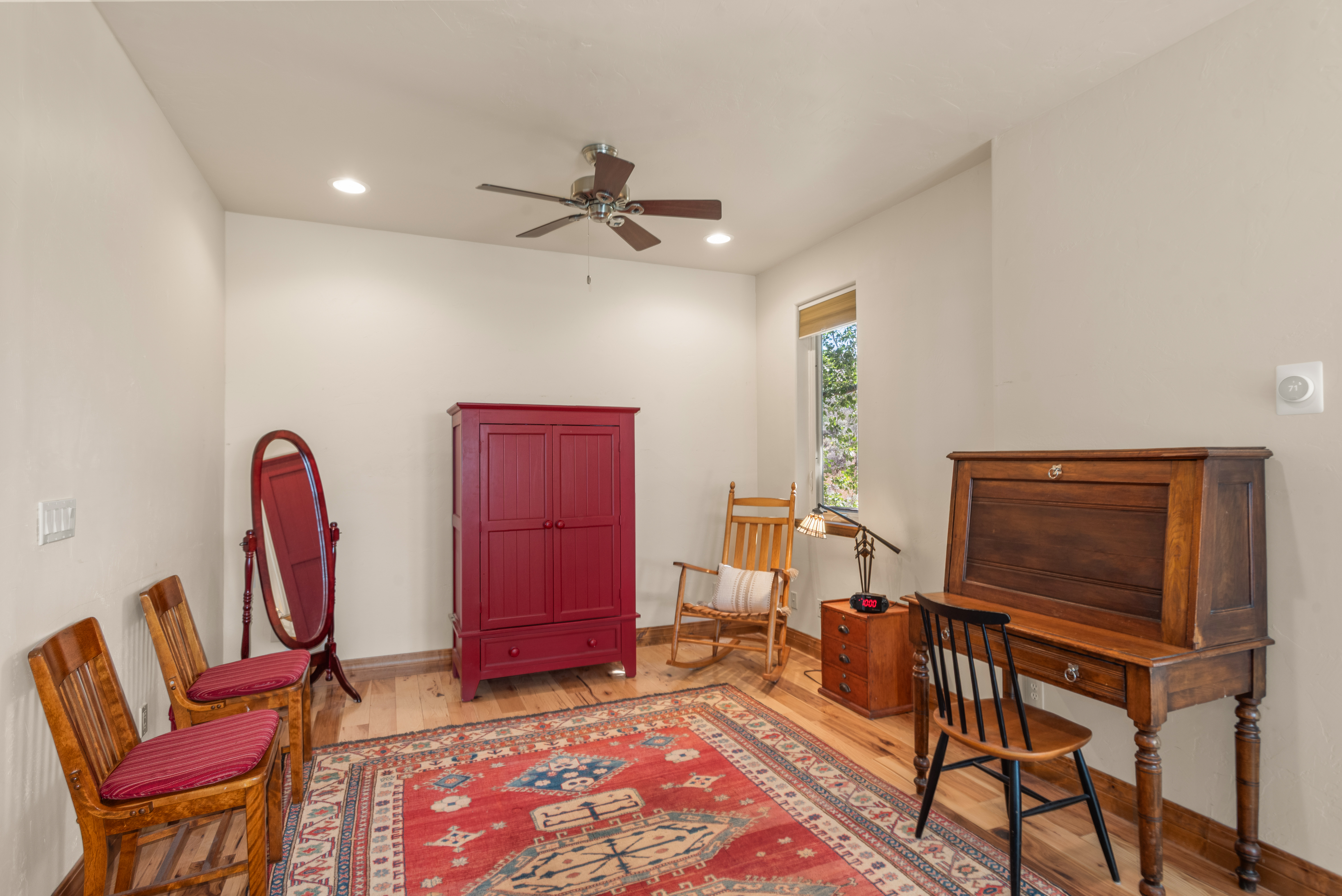 This study or multipurpose room features warm wood flooring, a striking red armoire, and a classic roll-top writing desk. The space is accented by a vibrant, traditional-style area rug, a wooden rocking chair, and an oval floor mirror. It provides a cozy and functional atmosphere with a nice blend of antique-inspired furniture.