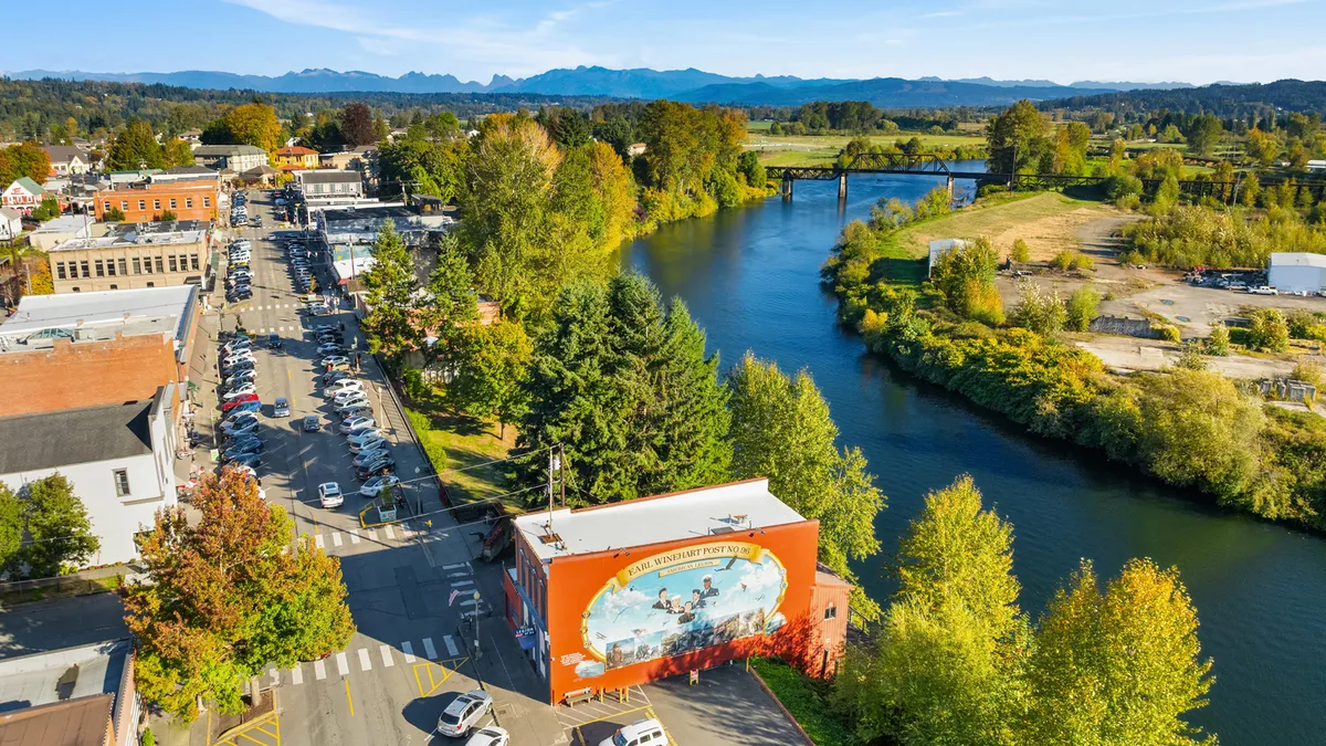 This aerial shot showcases the town's charm, featuring a river running alongside the main street lined with parked cars. The building with a large mural adds a unique artistic touch. The surrounding trees and distant mountains create a scenic and inviting atmosphere, highlighting the area's natural beauty.