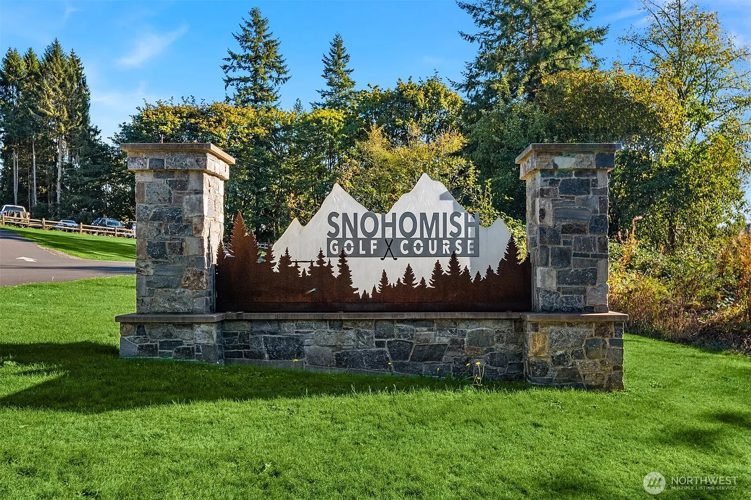 The image showcases the entrance to the Snohomish Golf Course, featuring an artistic sign mounted on a stone base between two stone pillars. The sign prominently displays the golf course's name against a mountain silhouette, with stylized trees below. The well-maintained green lawn in the foreground contributes to a welcoming and aesthetically pleasing first impression.