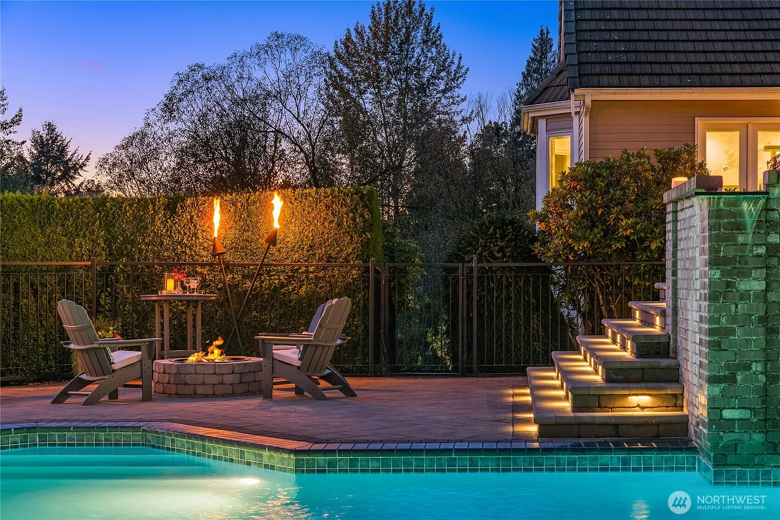 This image showcases a luxurious outdoor pool and patio area, perfect for relaxation and entertainment. The scene features a well-maintained pool with tile accents, a stone fire pit surrounded by Adirondack chairs, and decorative tiki torches. Steps with integrated lighting lead to an elevated section, enhancing the ambiance and visual appeal of this backyard oasis.