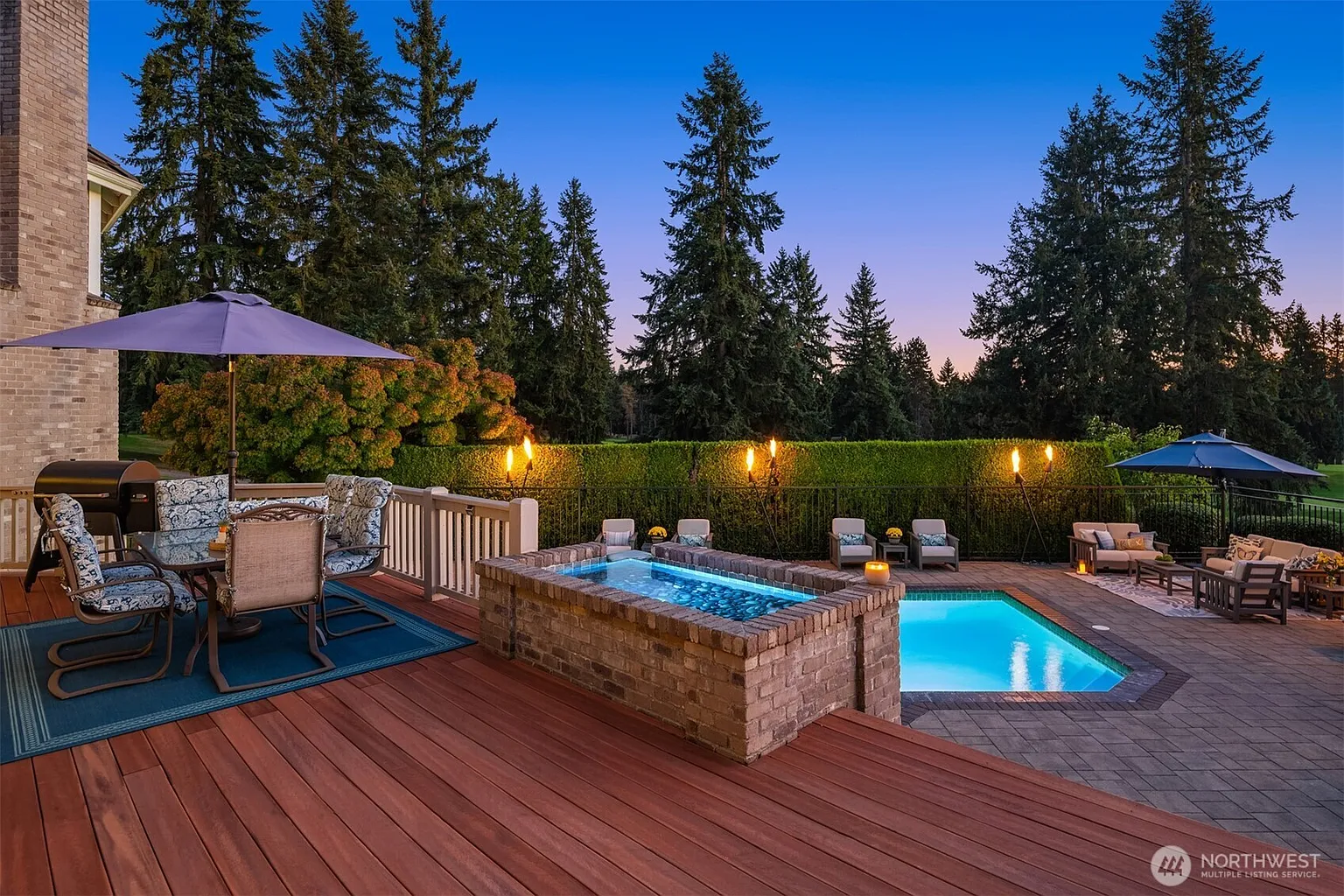 This image showcases an inviting outdoor living space complete with a pool and spa. A wooden deck area features patio furniture and a grill, while the pool and spa are surrounded by brick and stone. Mature trees and landscaping create a private and serene backyard setting.