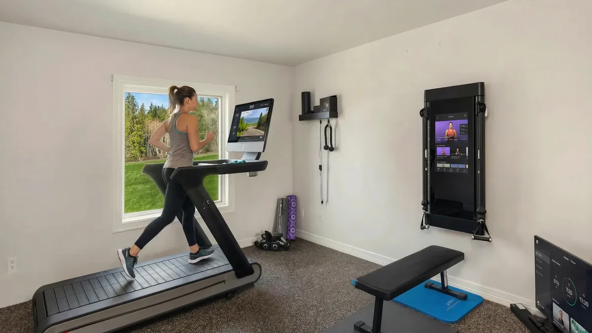 This is a well-equipped home gym with a treadmill, a Tonal system mounted on the wall, and a weight bench. The room features neutral paint and carpet, creating a functional and inviting workout space. Natural light from a window adds to the appeal.