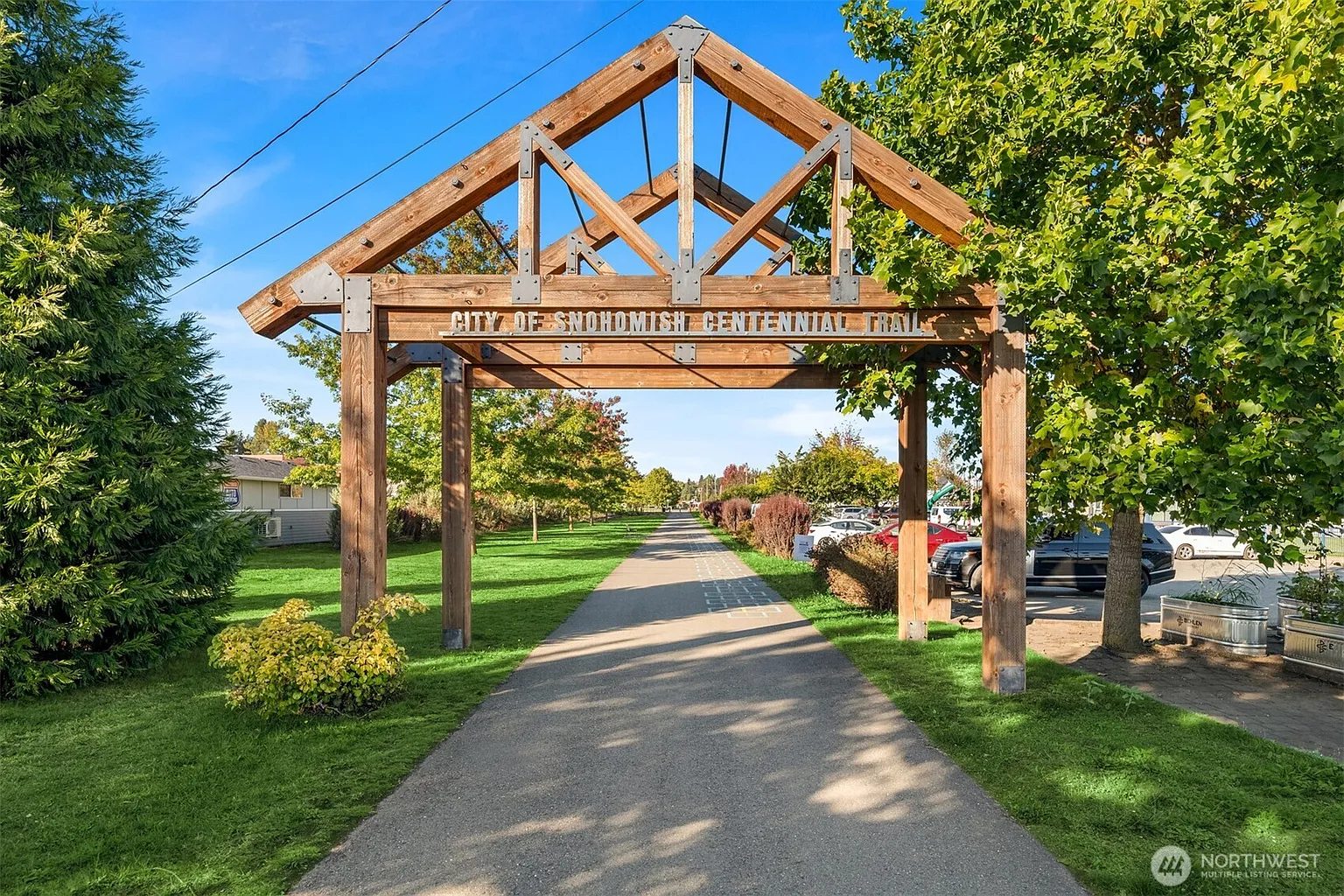 This image showcases the entrance to the "City of Snohomish Centennial Trail" through a wooden archway. The trail extends into the distance, flanked by green lawns and mature trees. The scene is well-maintained and inviting, suggesting a pleasant outdoor recreational area.