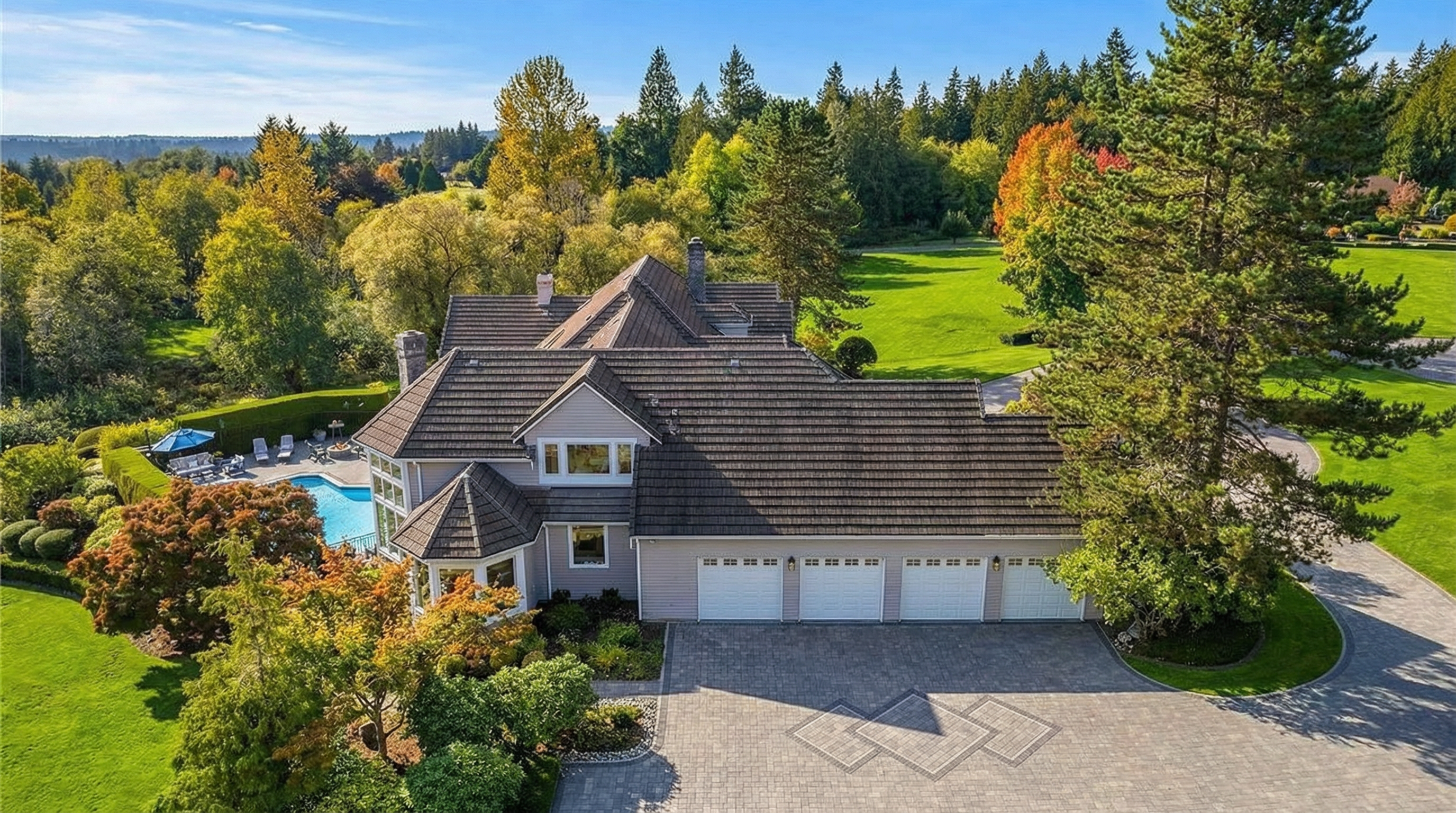 This aerial view showcases a luxurious residential property with well-manicured grounds. The house features a prominent roof, multiple garage doors, and a nearby swimming pool. Mature trees and lawns surround the home, creating a private and upscale estate setting.