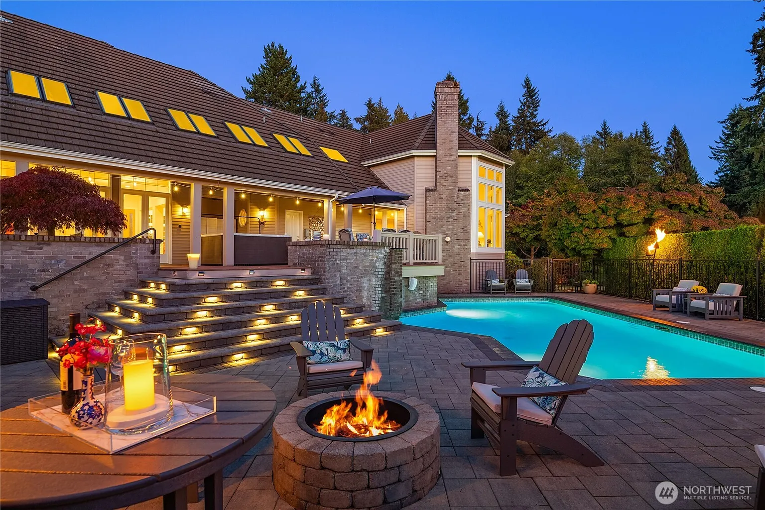 This image showcases a luxurious backyard pool and spa area. A stone patio surrounds the pool, featuring lounge chairs and a fire pit. The well-lit steps lead to an upper deck with a spa, blending seamlessly with the home's architecture and lush landscaping, which creates a resort-like ambiance.