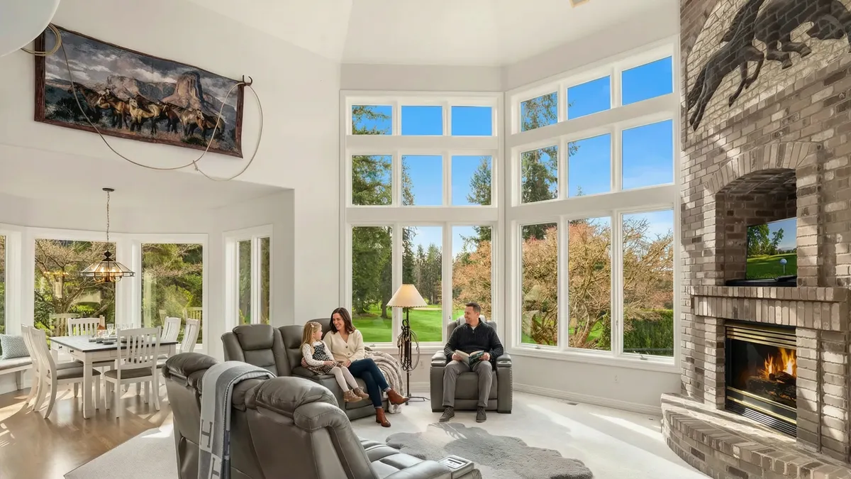 The image showcases a bright and spacious living room with tall windows offering picturesque outdoor views. Natural light floods the room, highlighting the neutral color palette and comfortable seating arrangement. A brick fireplace with a mounted TV adds a focal point, while the overall atmosphere exudes comfort and modern elegance.