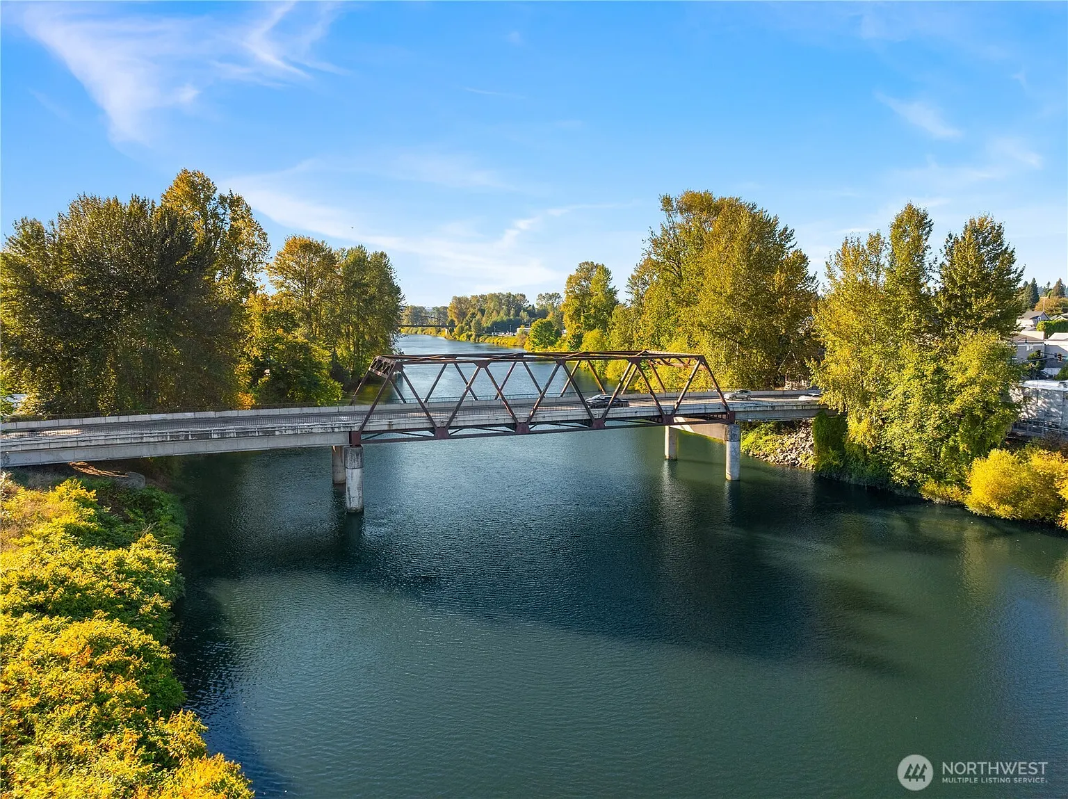 This aerial shot showcases a bridge over a river, surrounded by lush green trees under a clear blue sky. The river's surface reflects the sky, adding depth to the scene. The composition emphasizes the natural beauty of the surroundings, which would be attractive for a property listing focused on nature or river access.