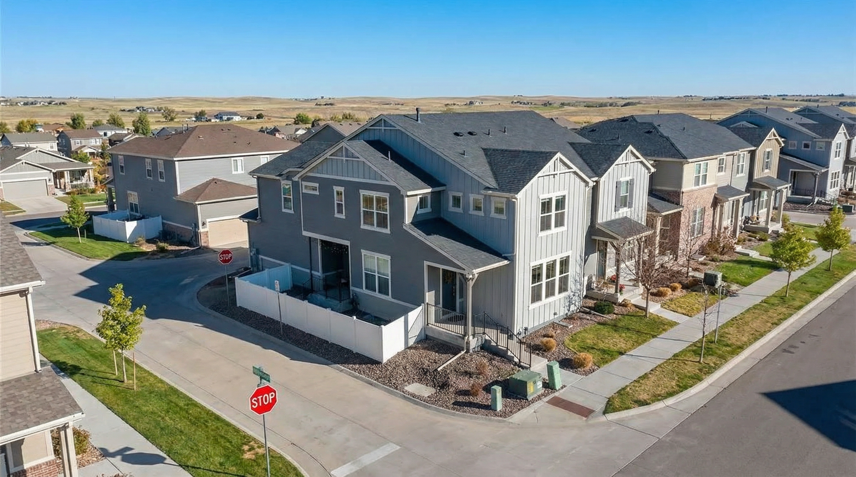 This is an aerial view showcasing a row of modern, two-story houses in a residential neighborhood. The homes exhibit varying shades of gray and neutral-toned exteriors, with dark gray roofing that contrasts with the siding. Manicured lawns, sidewalks, and trees line the streets, contributing to a clean and well-maintained community appearance.