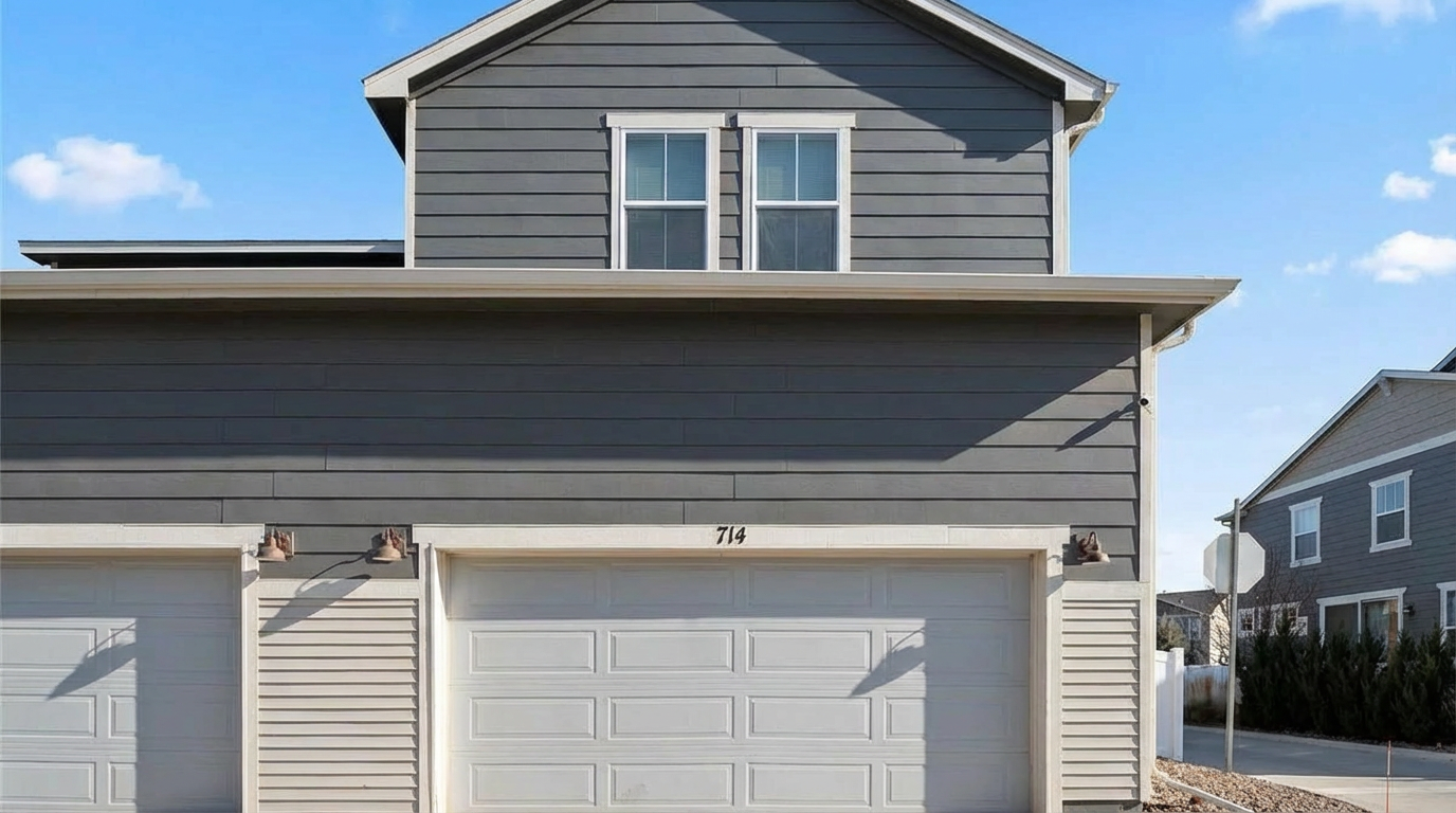 The image presents a close-up of a two-story home's garage facade, showcasing a double garage door and a single garage door. The garage doors are white with recessed panels, and the residence number '714' is prominently displayed above the main garage door. The exterior features horizontal gray siding and off-white vertical siding on the right side of the garage, accented by outdoor lighting fixtures.