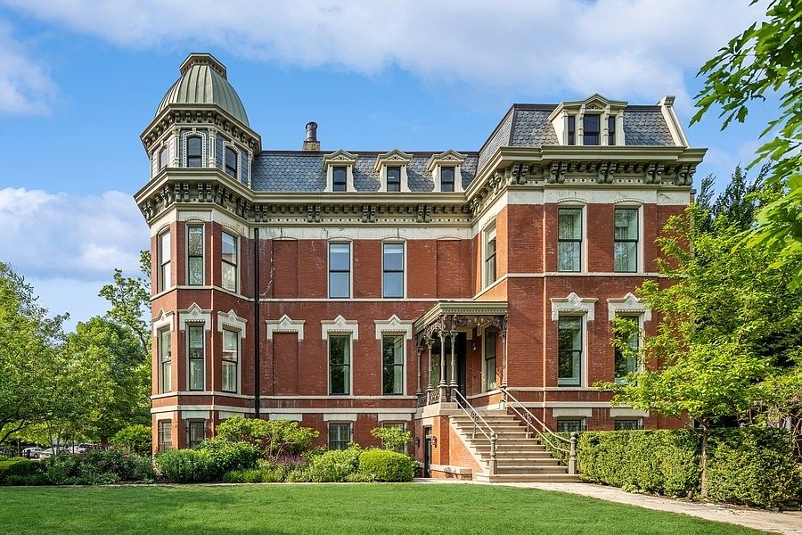 This grand, historic brick mansion features an iconic Second Empire architectural style, complete with a prominent corner tower capped by a mansard roof and intricate detailing. The stately facade is complemented by a covered front entryway with detailed ironwork and a sweeping staircase leading up to the main entrance. Surrounded by lush, manicured landscaping, the property presents a distinguished and elegant outward appearance.